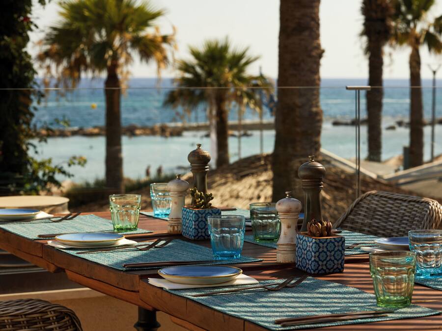 A dining table set with plates, glasses, and cutlery, overlooking a beach with palm trees and the ocean.