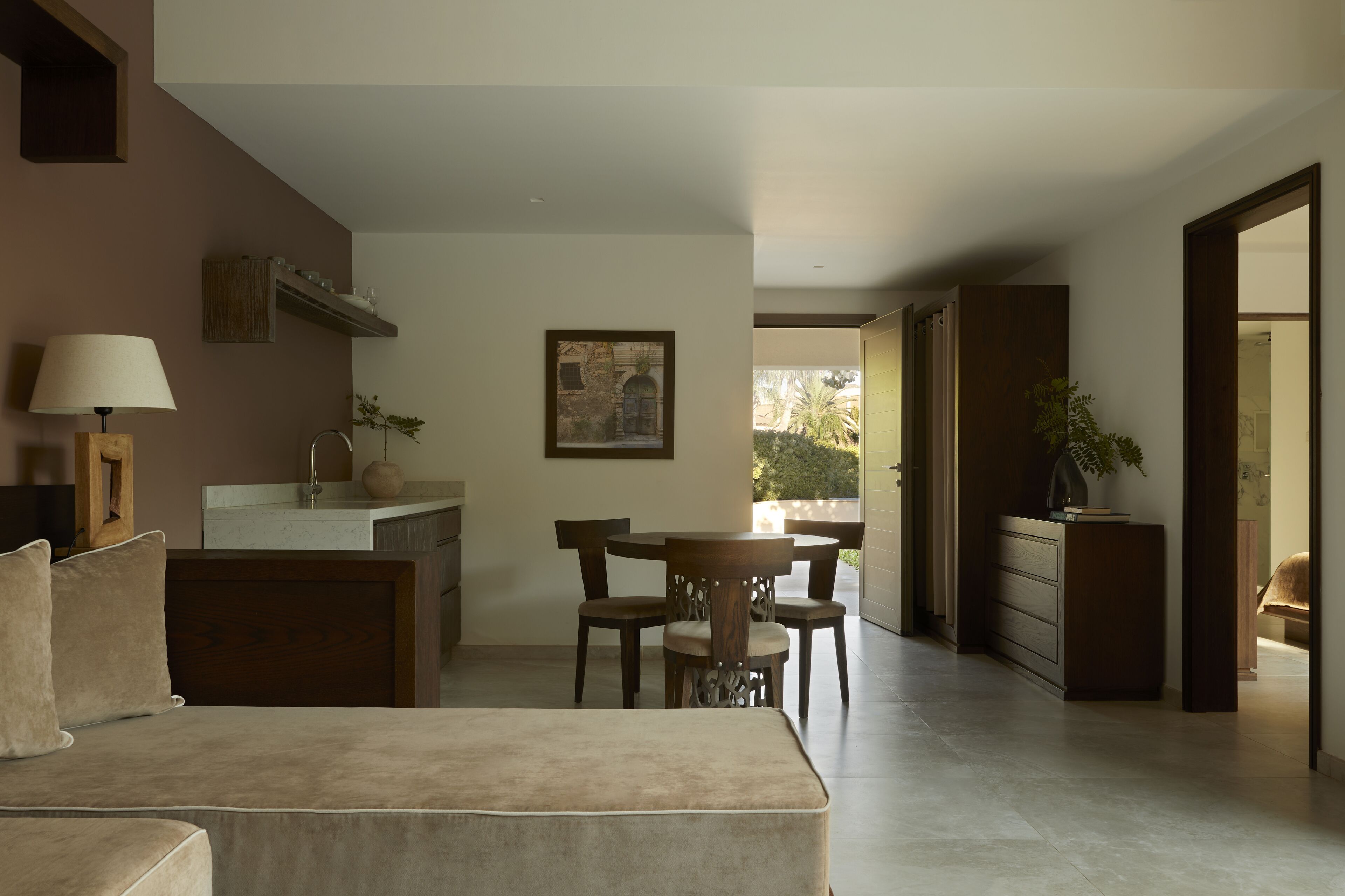 A modern living room with a sofa, dining table, and chairs. There is a kitchen area with a counter and cabinets. A framed picture hangs on the wall, and a plant is on the counter.