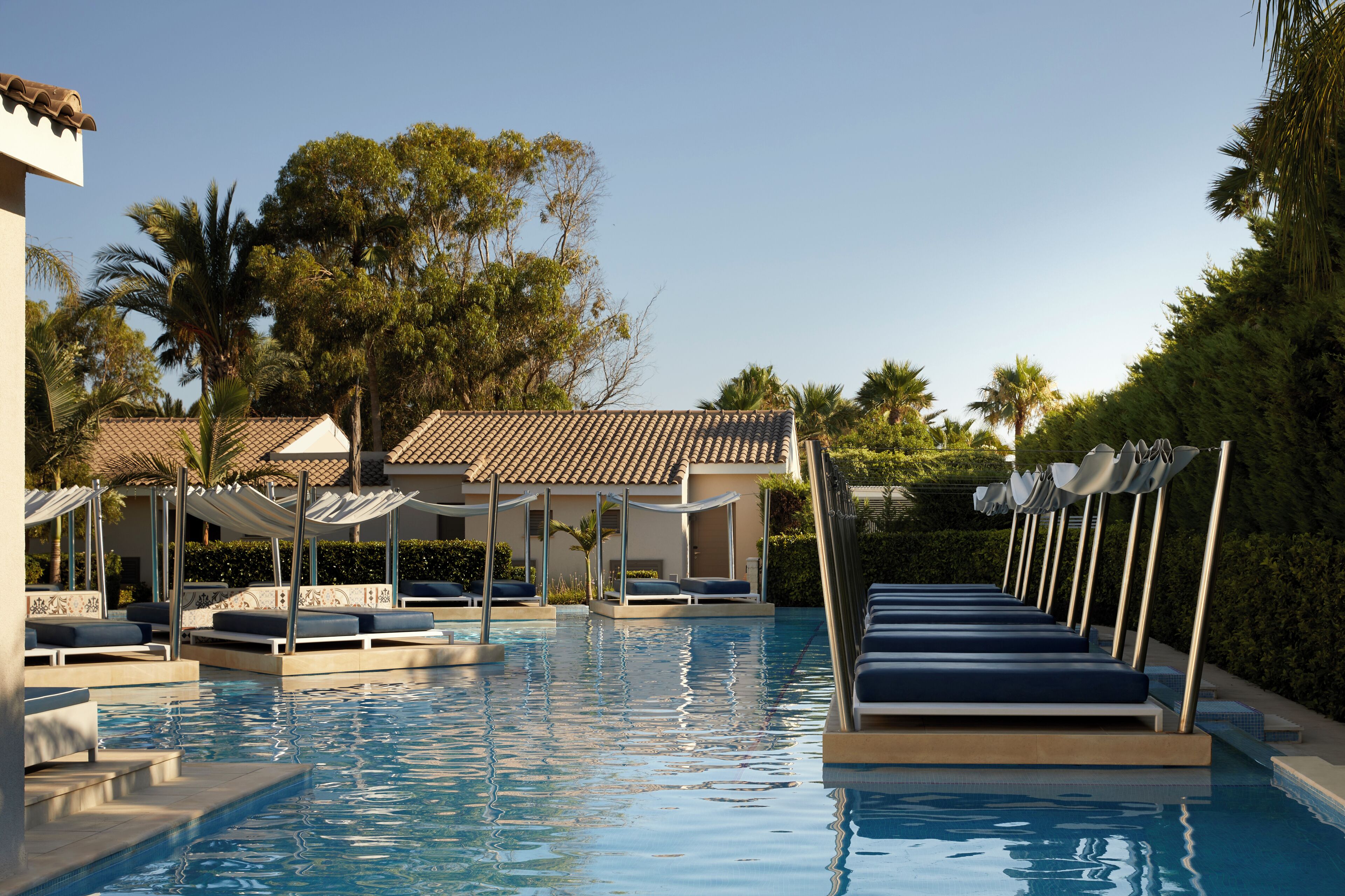 Una piscina con lettini, una zona relax coperta e un edificio con tetto in tegole sullo sfondo.