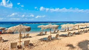 Een strand met rijen ligbedden en rieten parasols, een helderblauwe lucht en de oceaan op de achtergrond.
