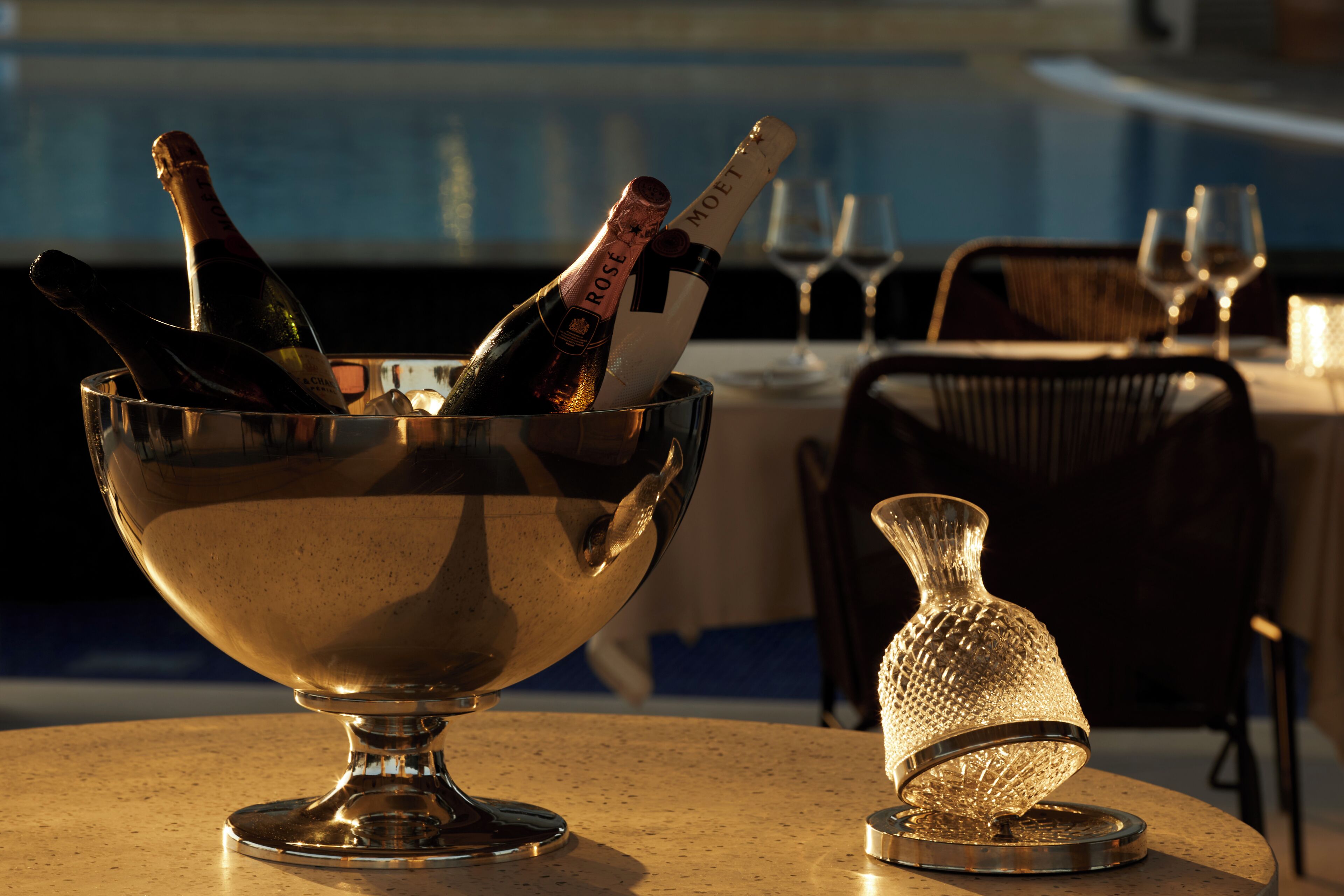 A bar area with a metal bowl containing champagne bottles, a glass vase, and a pool in the background.