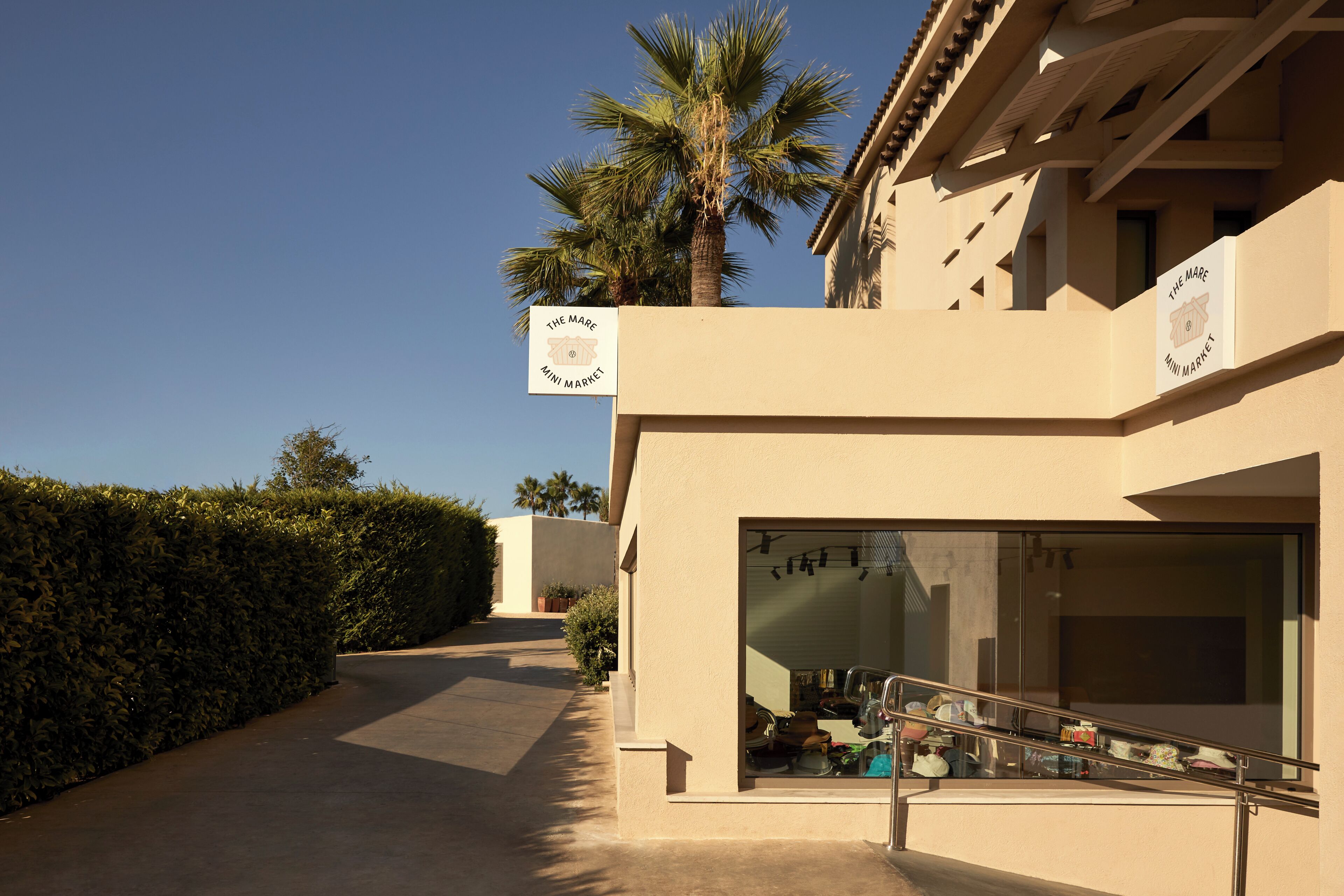 A building with a sign reading 'The Mare Mini Market,' a glass storefront, and a palm tree in the background.