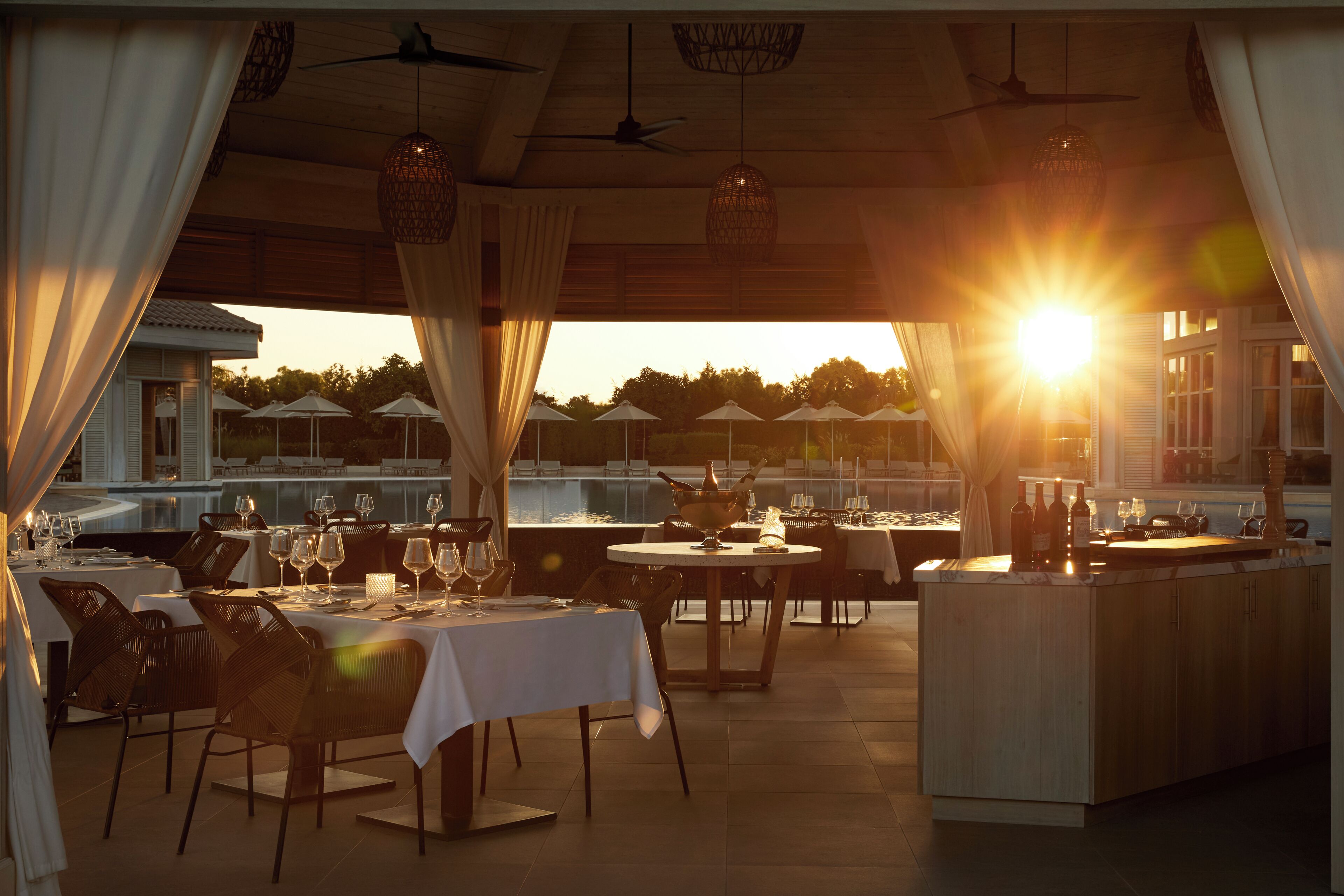 An outdoor dining area with tables set for two, a bar area, and a view of the sunset.