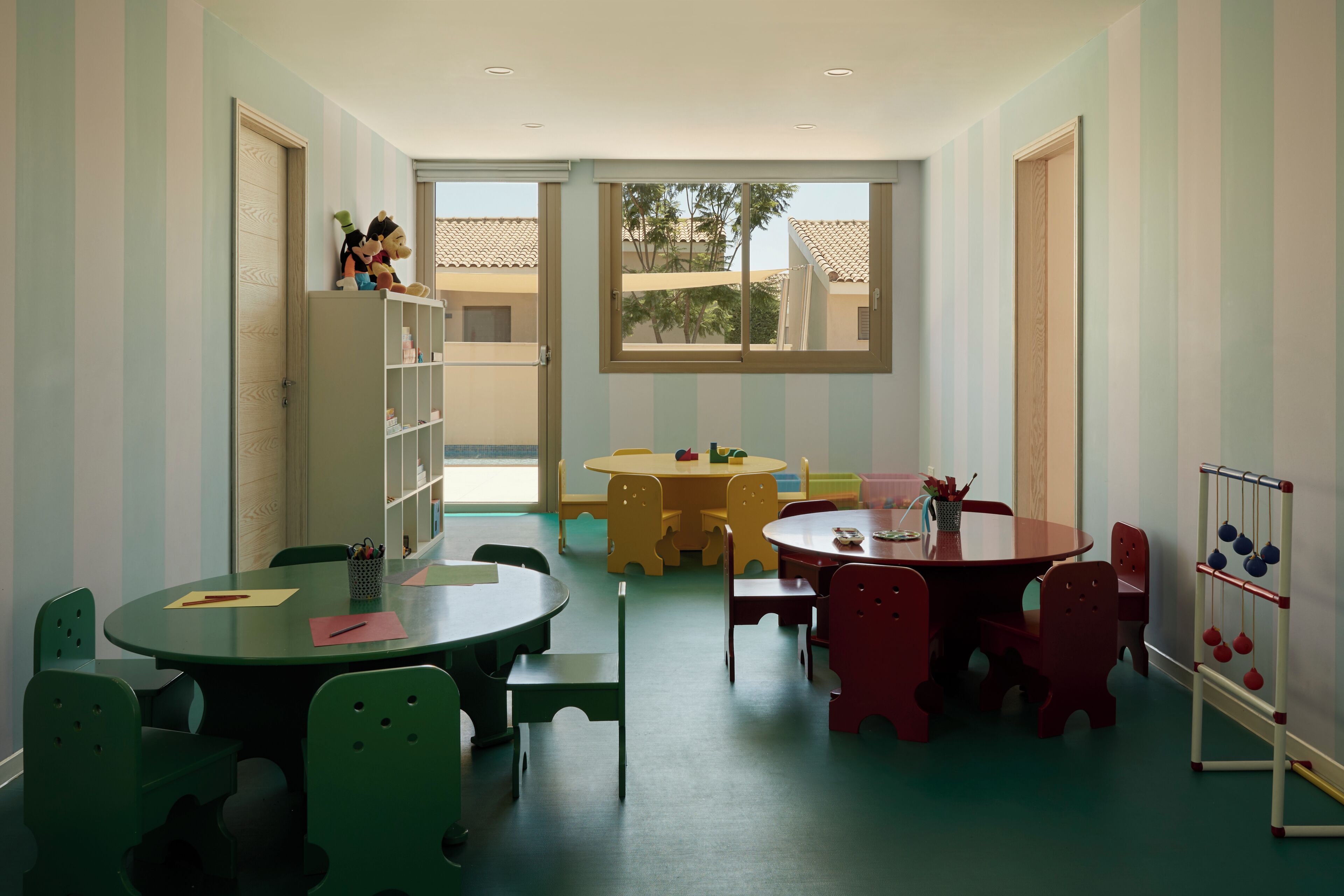 A children's playroom with striped walls, multiple tables, chairs, and a toy rack.