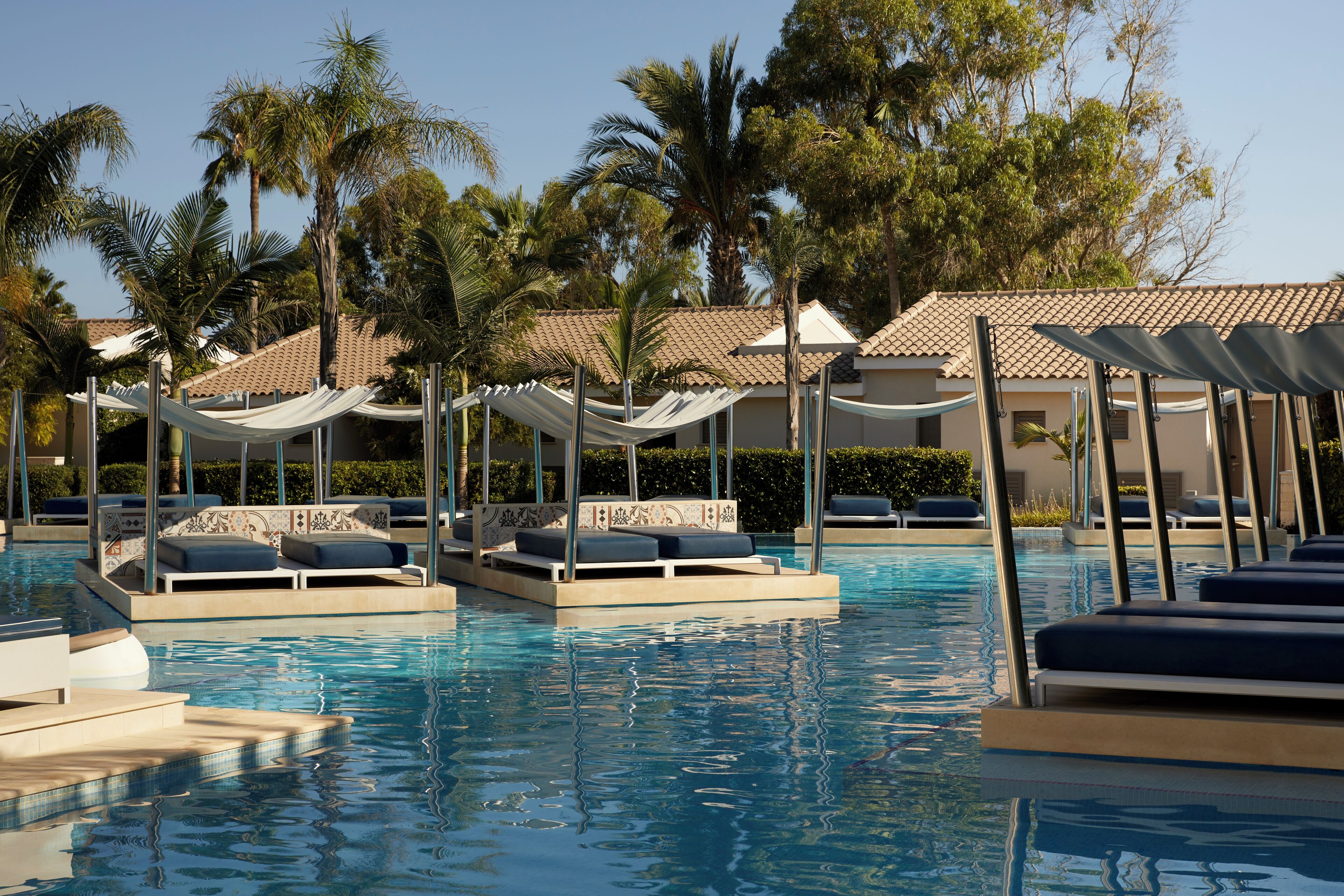 Zona de piscina con sillones de descanso y sombrillas, rodeada de palmeras y edificios con techos de tejas.