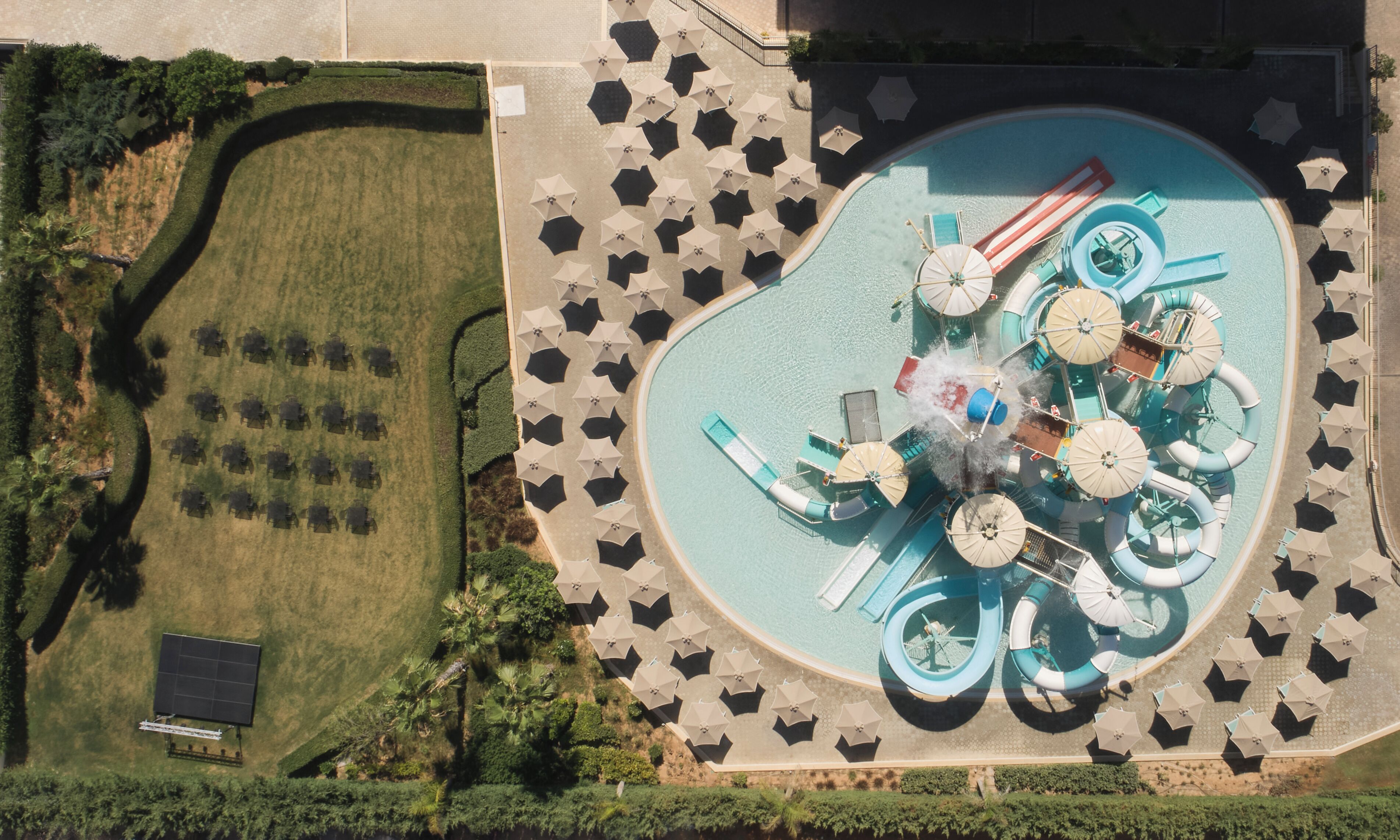 An aerial view of a resort with a large pool, multiple water slides, and numerous umbrellas.