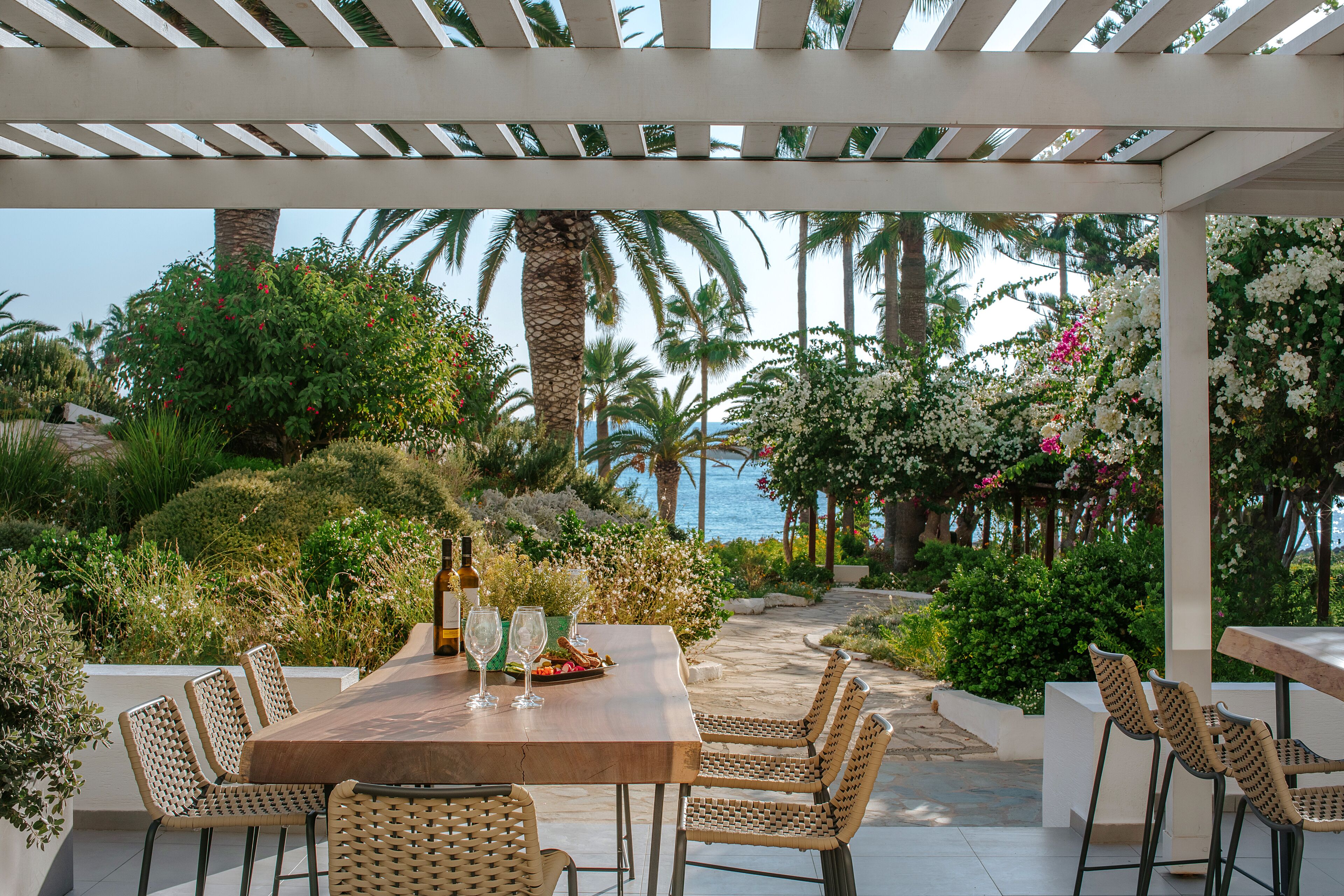 Une terrasse extérieure avec une table en bois et des chaises en osier, offrant une vue sur un jardin luxuriant et la mer.