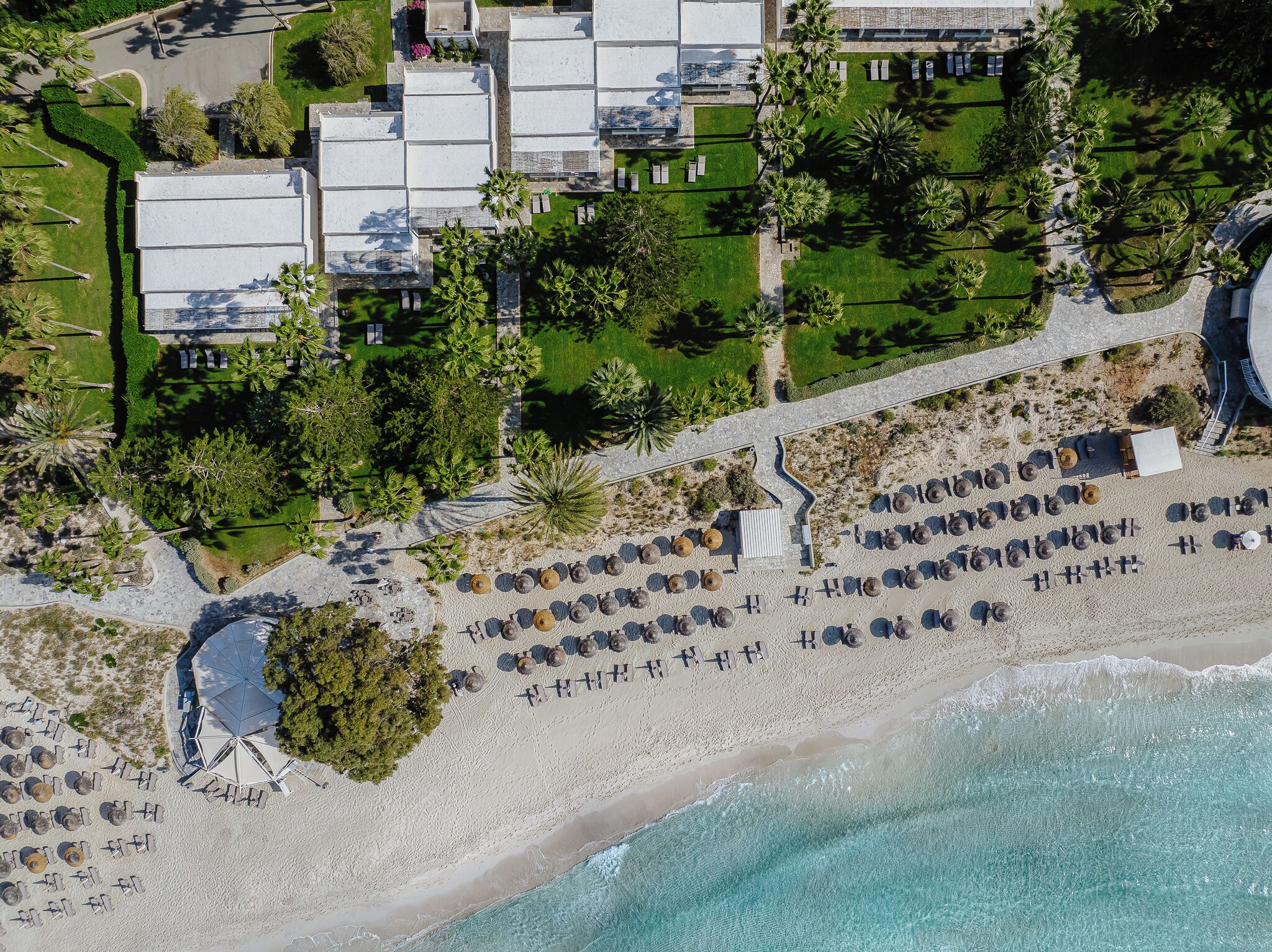 An aerial view of a resort with multiple buildings, a beach with rows of sun loungers, and palm trees.
