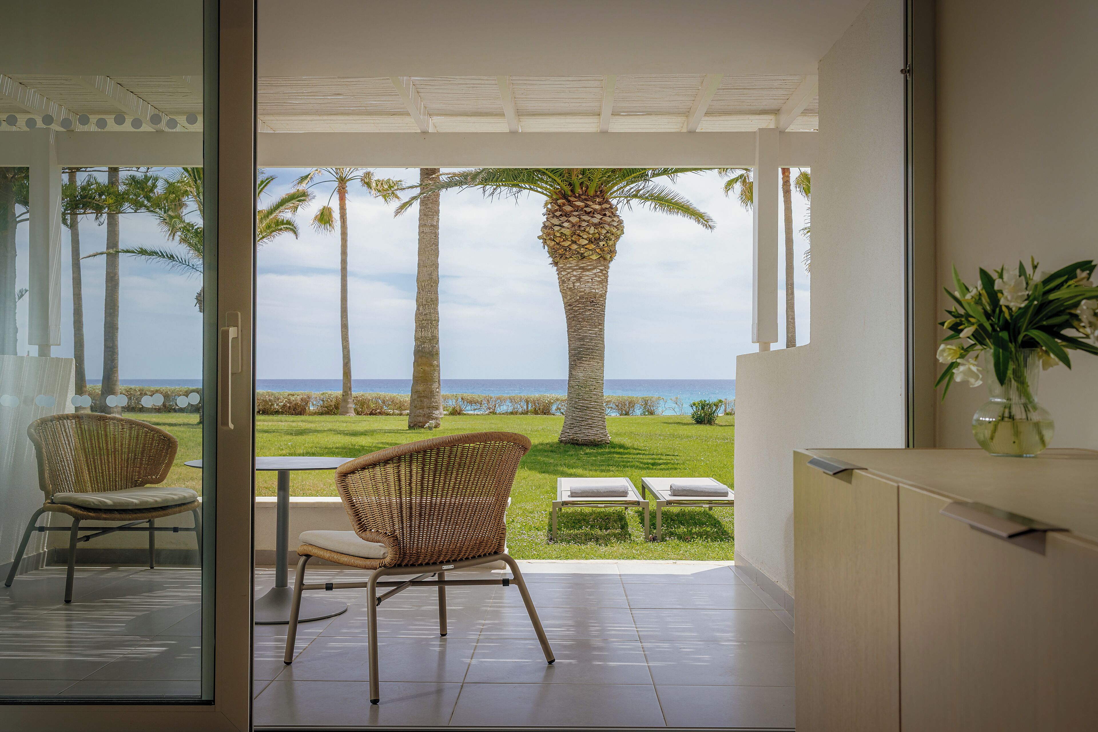 Una habitación con puerta de cristal que da a un balcón con sillas de mimbre y vistas a palmeras y al mar.