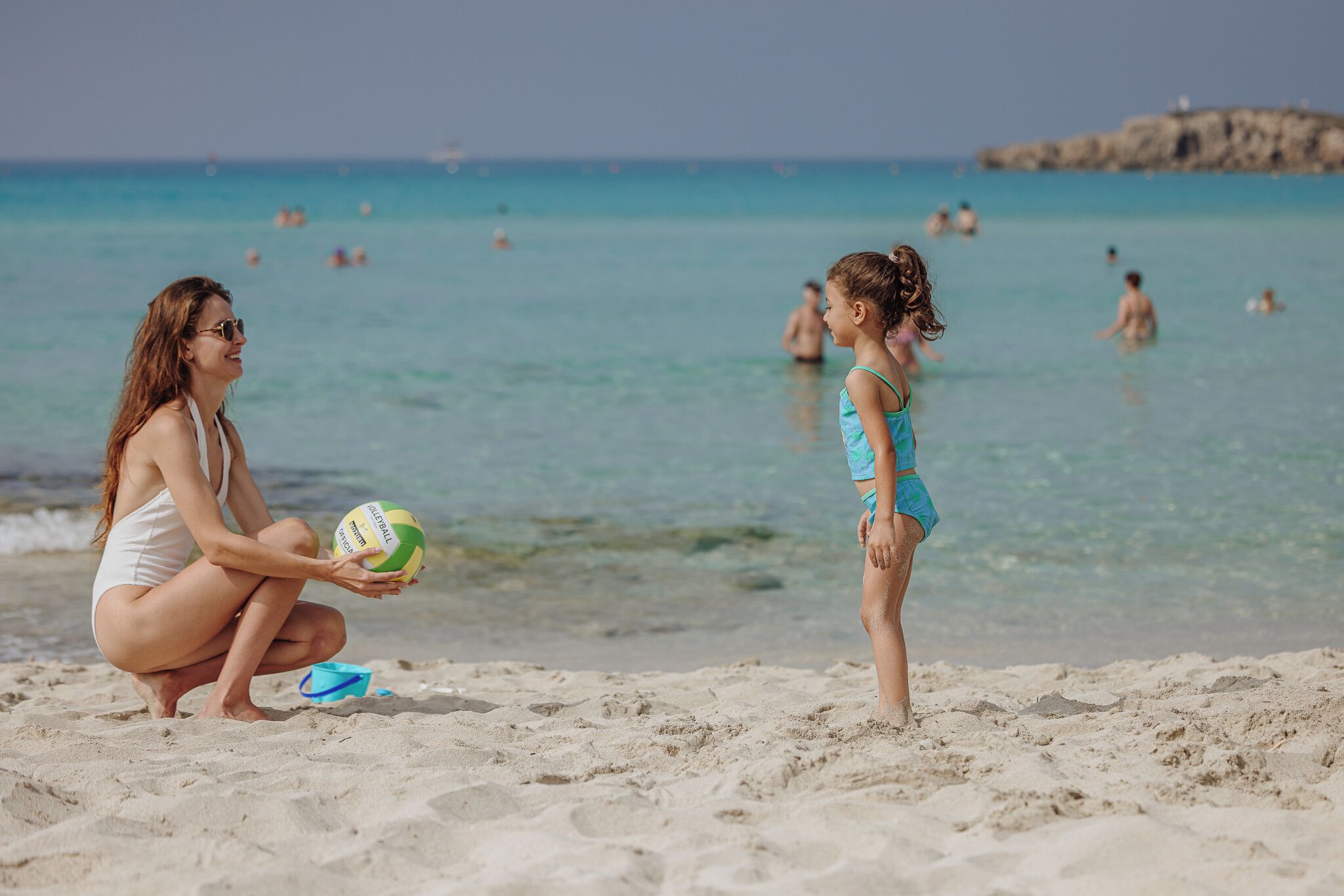 A beach scene with a woman and a child playing with a volleyball.