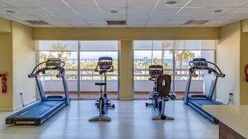 Ett gym med löpband och crosstrainers, stora fönster med utsikt över stranden och palmerna, och en brandsläckare på väggen.
