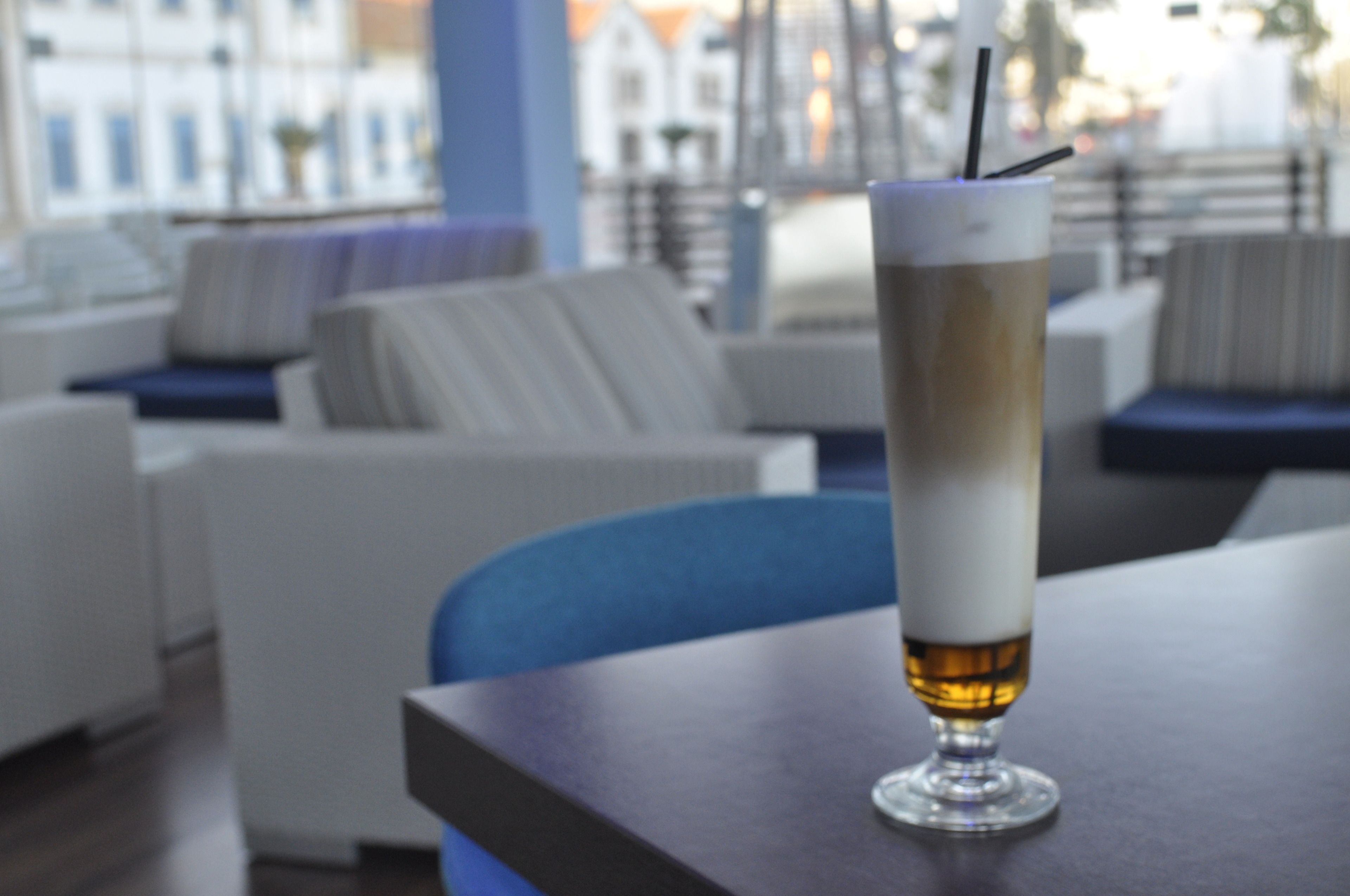 A tall glass of beer on a table in a modern lounge area with white sofas and a view of buildings in the background.