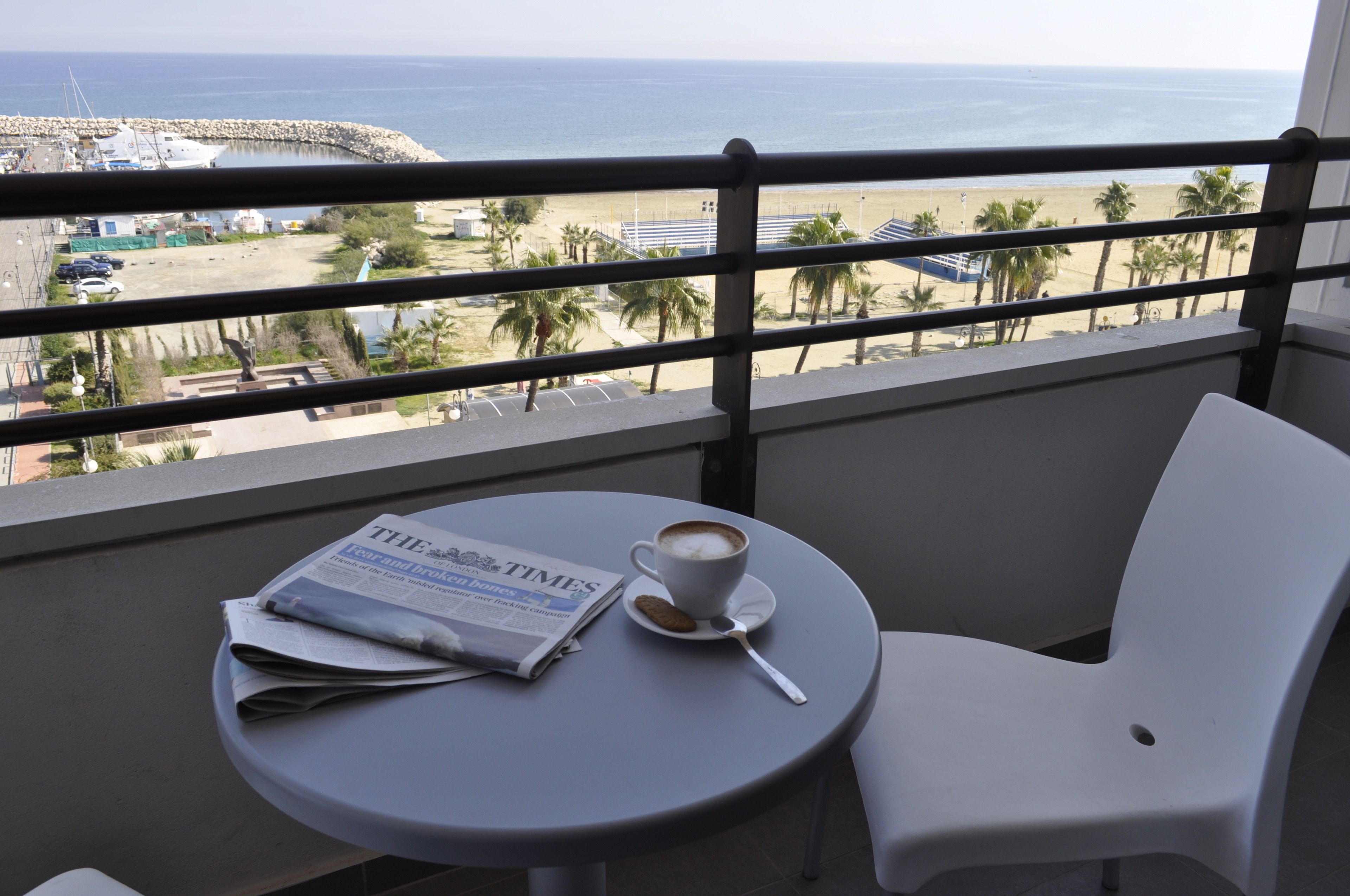Un balcón con una mesa, una silla, un periódico, una taza de café y vistas a la playa y al océano.