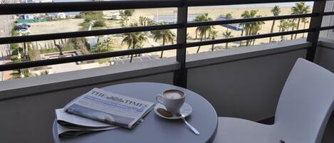 A balcony with a table, a chair, a newspaper, a cup of coffee, and a view of the beach and ocean.