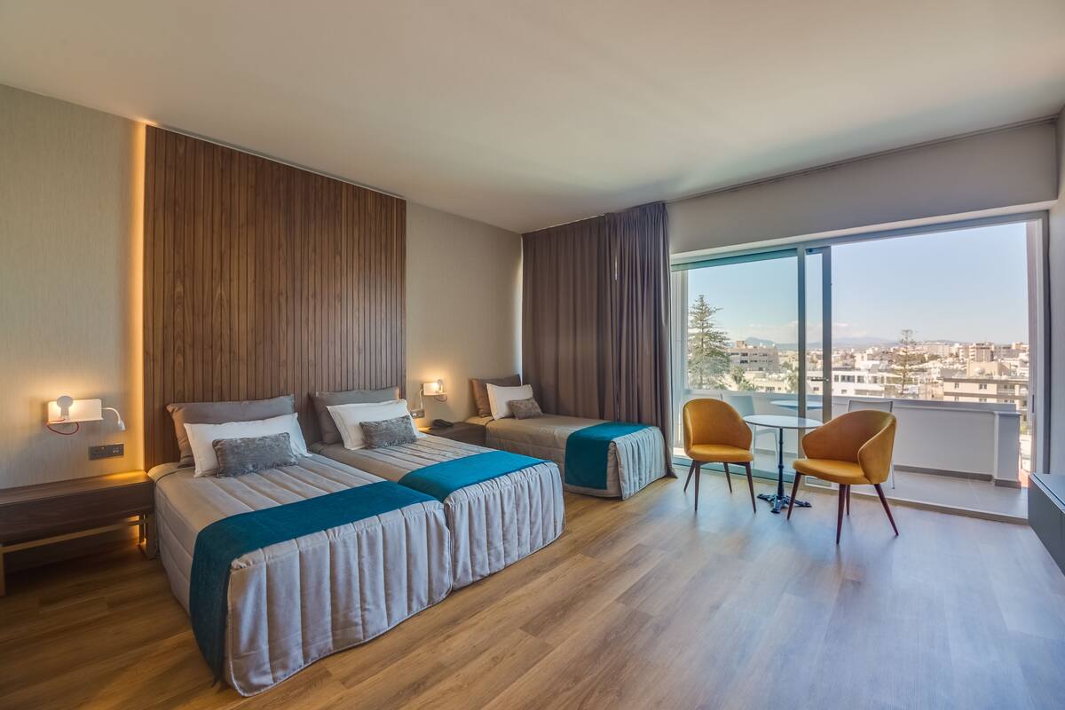 A modern hotel room with two beds, a balcony with a view, and a small table with chairs.