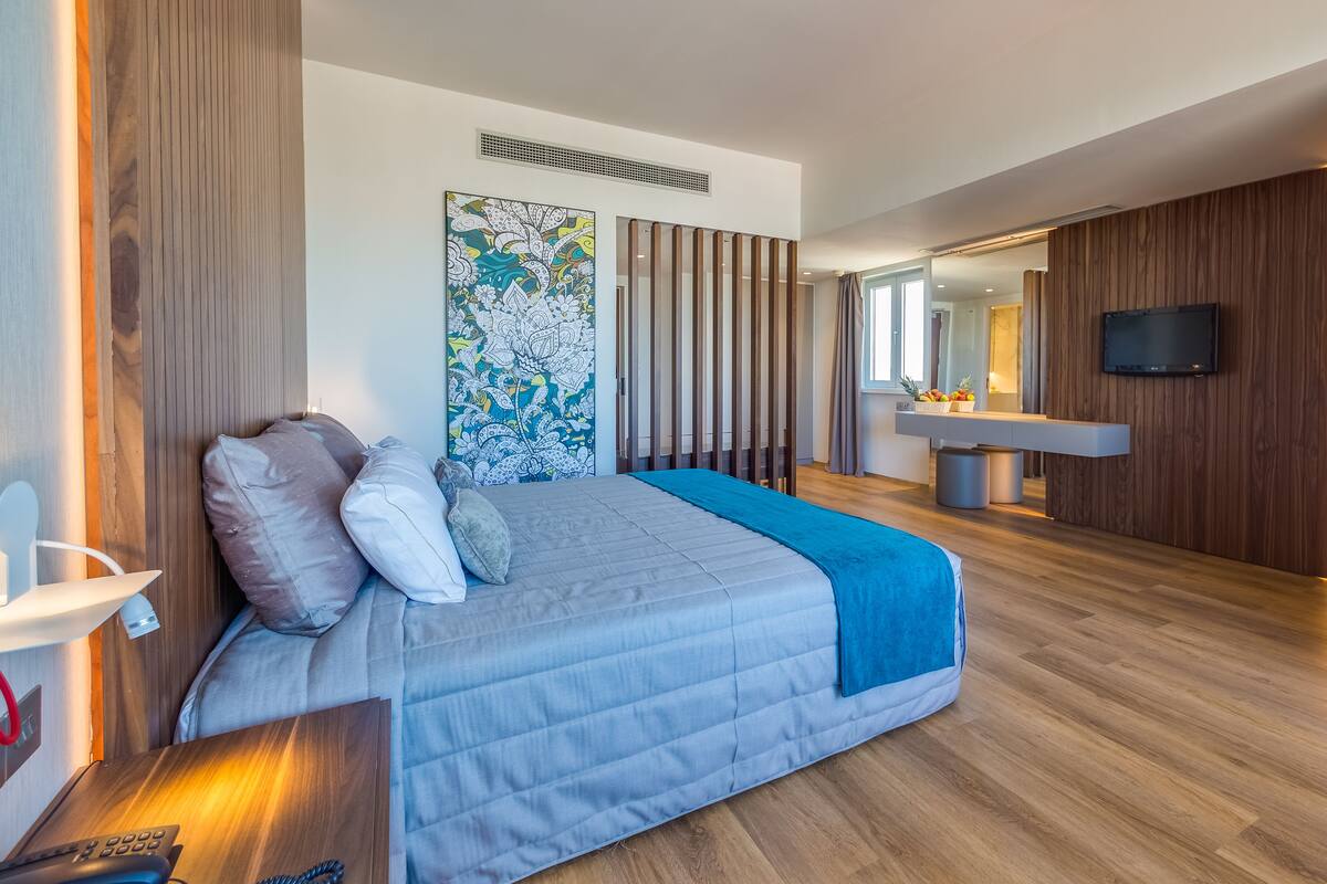 A modern bedroom with a large bed, a bedside table, a flat-screen TV mounted on the wall, and a wooden floor.