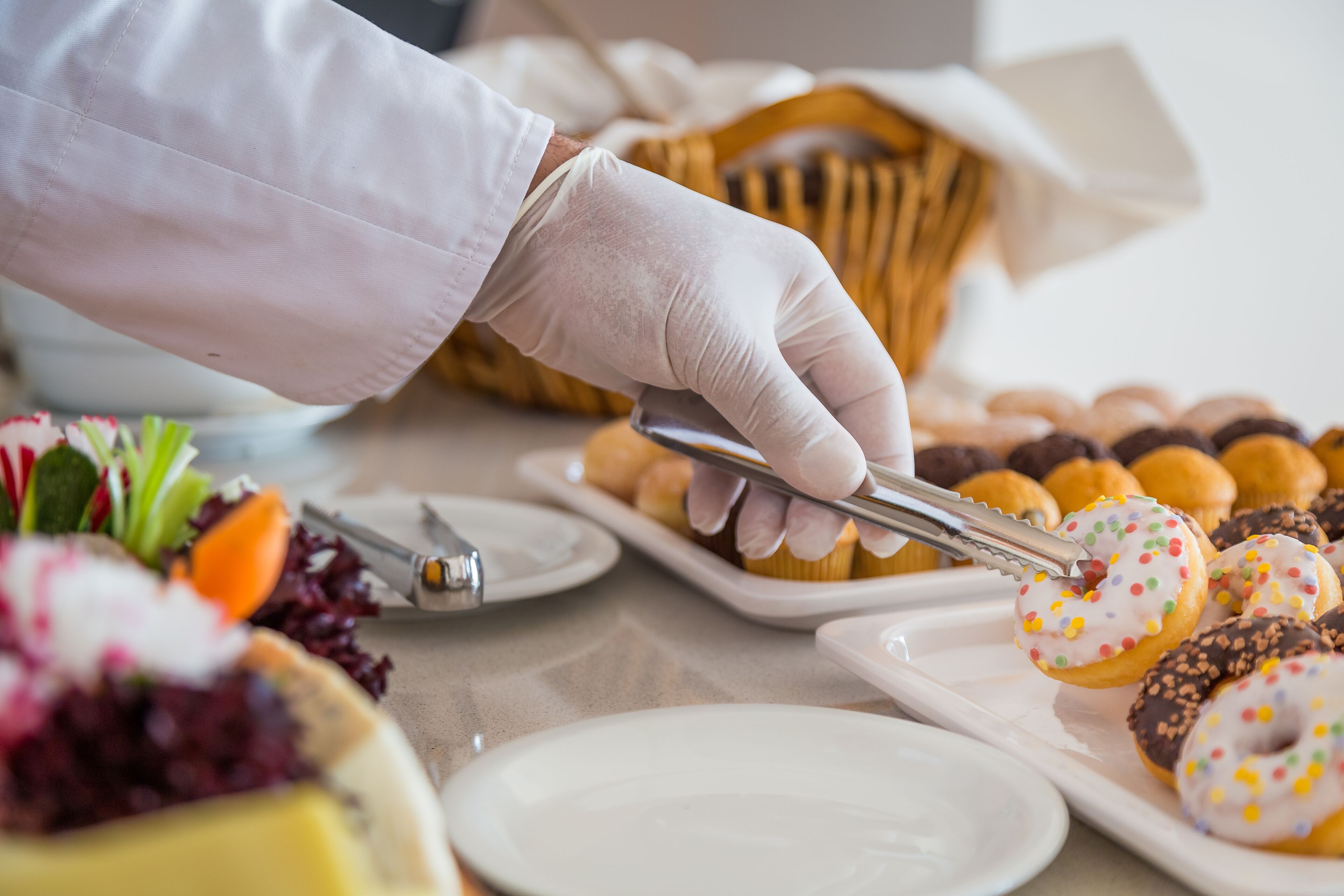 A person in a white glove handling food items, including cupcakes and pastries, on a table with various dishes and a basket in the background.