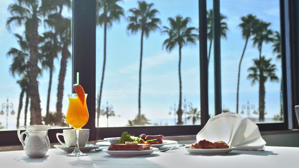 A table set with breakfast items, a glass of orange juice, and a view of palm trees and the ocean through large windows. - Sun Hall Hotel (Larnaca)
