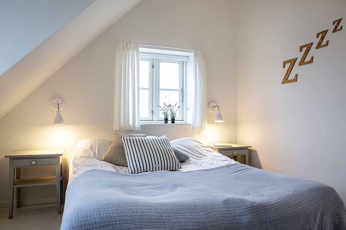 Une chambre avec un plafond en pente, un lit avec un oreiller à rayures, deux lampes de chevet et une fenêtre avec des rideaux blancs.