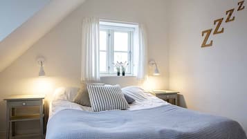 Un dormitorio con techo inclinado, una cama con almohada a rayas, dos lámparas de noche y un ventanal con cortinas blancas.