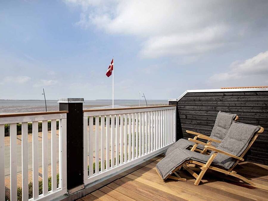 A wooden deck with a white railing, two wooden lounge chairs, and a view of the beach and ocean.