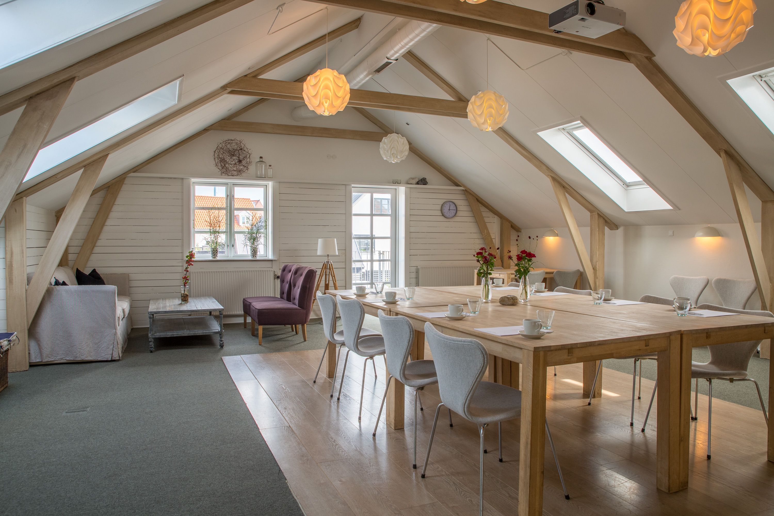 A spacious attic room with a high ceiling, wooden beams, skylights, a dining table with chairs, a sofa, and a small side table.