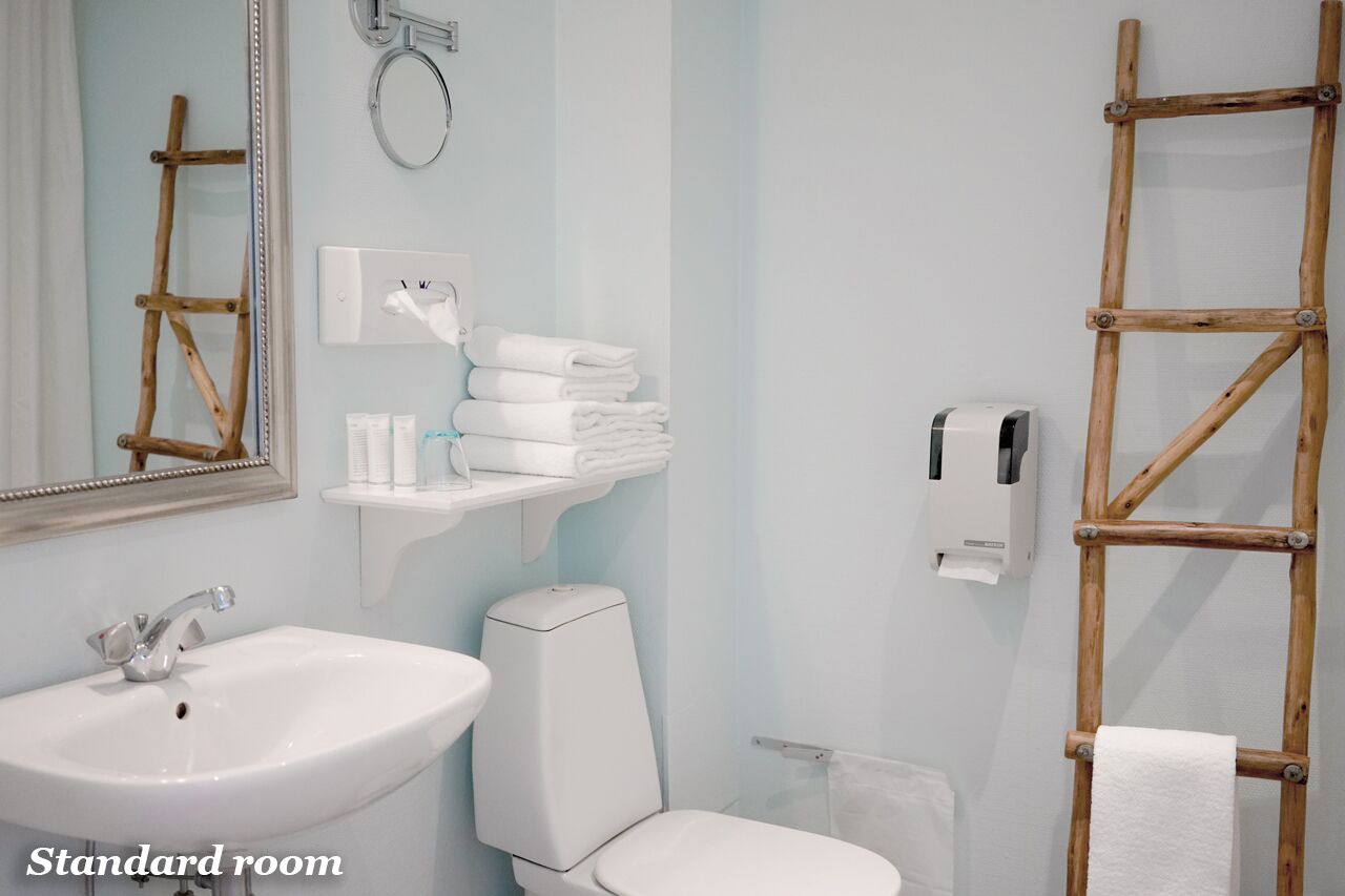 Et standardværelse med en hvid vask, toilet og et spejl. Der er håndklæder stablet på en hylde, en toiletpapirholder og en træstige.