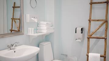 Et standardværelse med en hvid vask, toilet og et spejl. Der er håndklæder stablet på en hylde, en toiletpapirholder og en træstige.