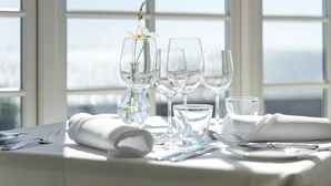 A table set for dining with wine glasses, a water glass, and cutlery, with a vase of flowers and a view of the sea through large windows. - Hjerting Badehotel - Esbjerg (Esbjerg)