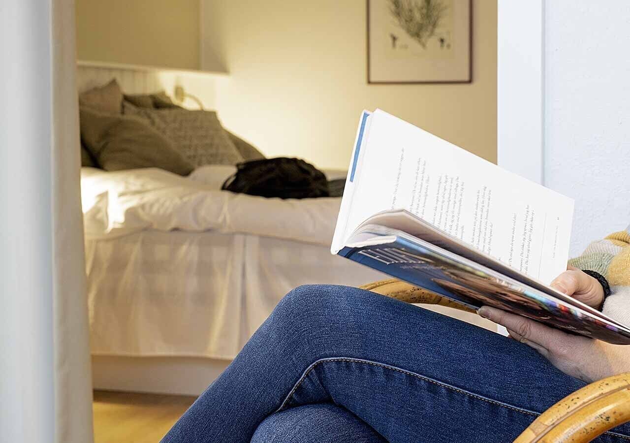 Iemand zit in een kamer op een stoel, een boek lezend, met een bed en een lijst aan de muur.