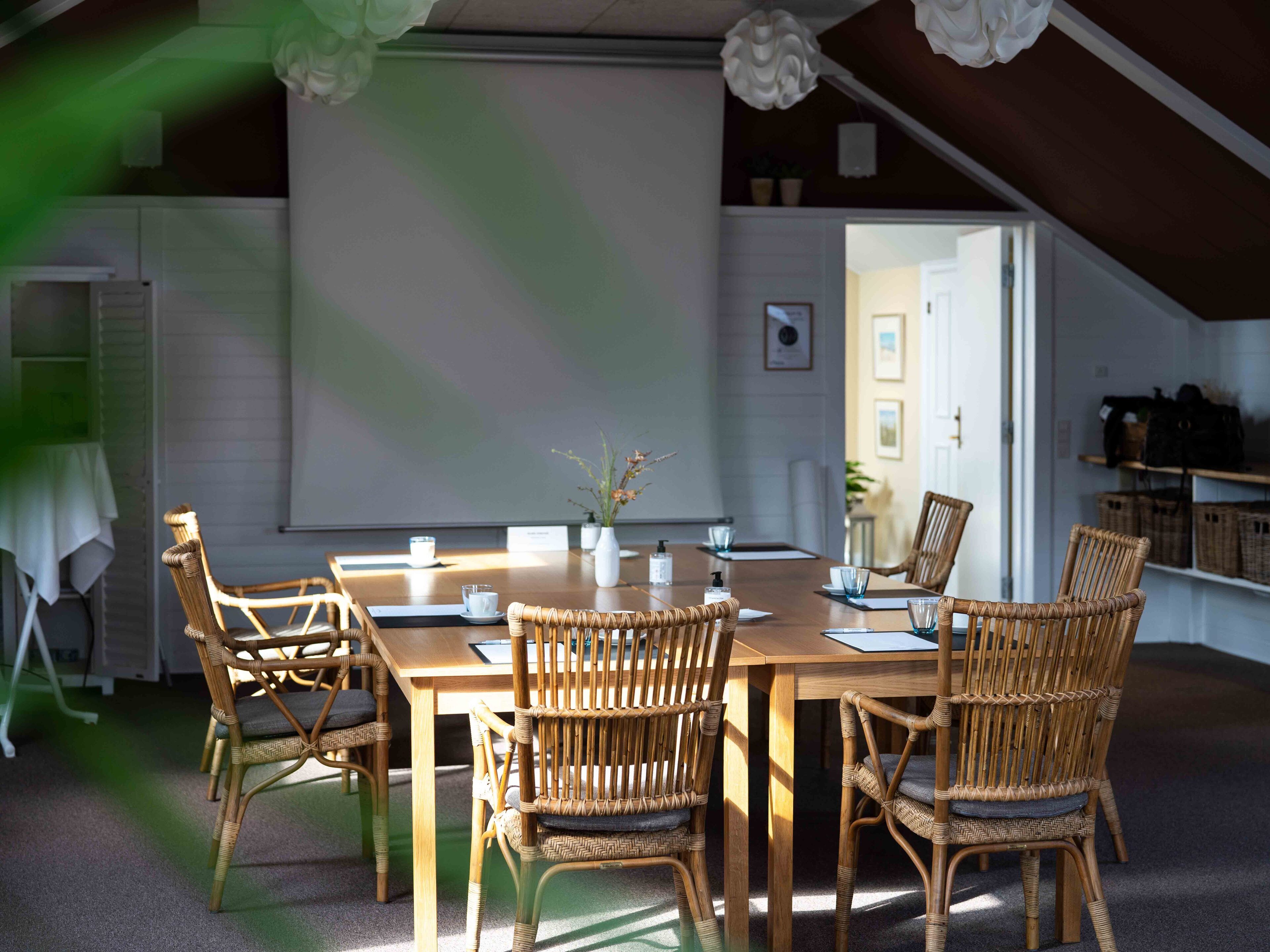 A meeting room with a large table, wicker chairs, a projector screen, and a door leading to another room.