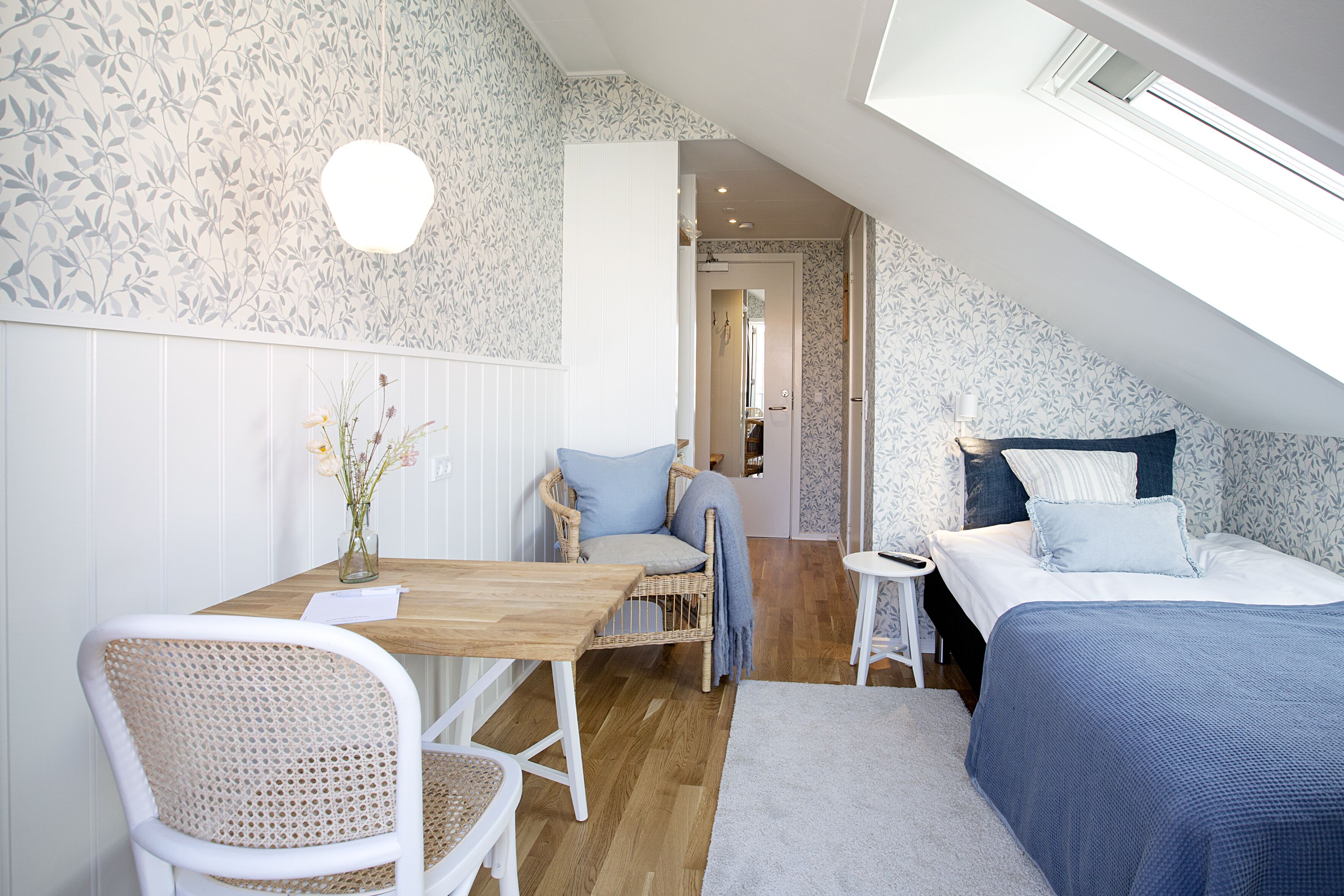 Una camera da letto con soffitto a falde, un letto con biancheria azzurra, un comodino, una sedia e un tavolino con un vaso di fiori.