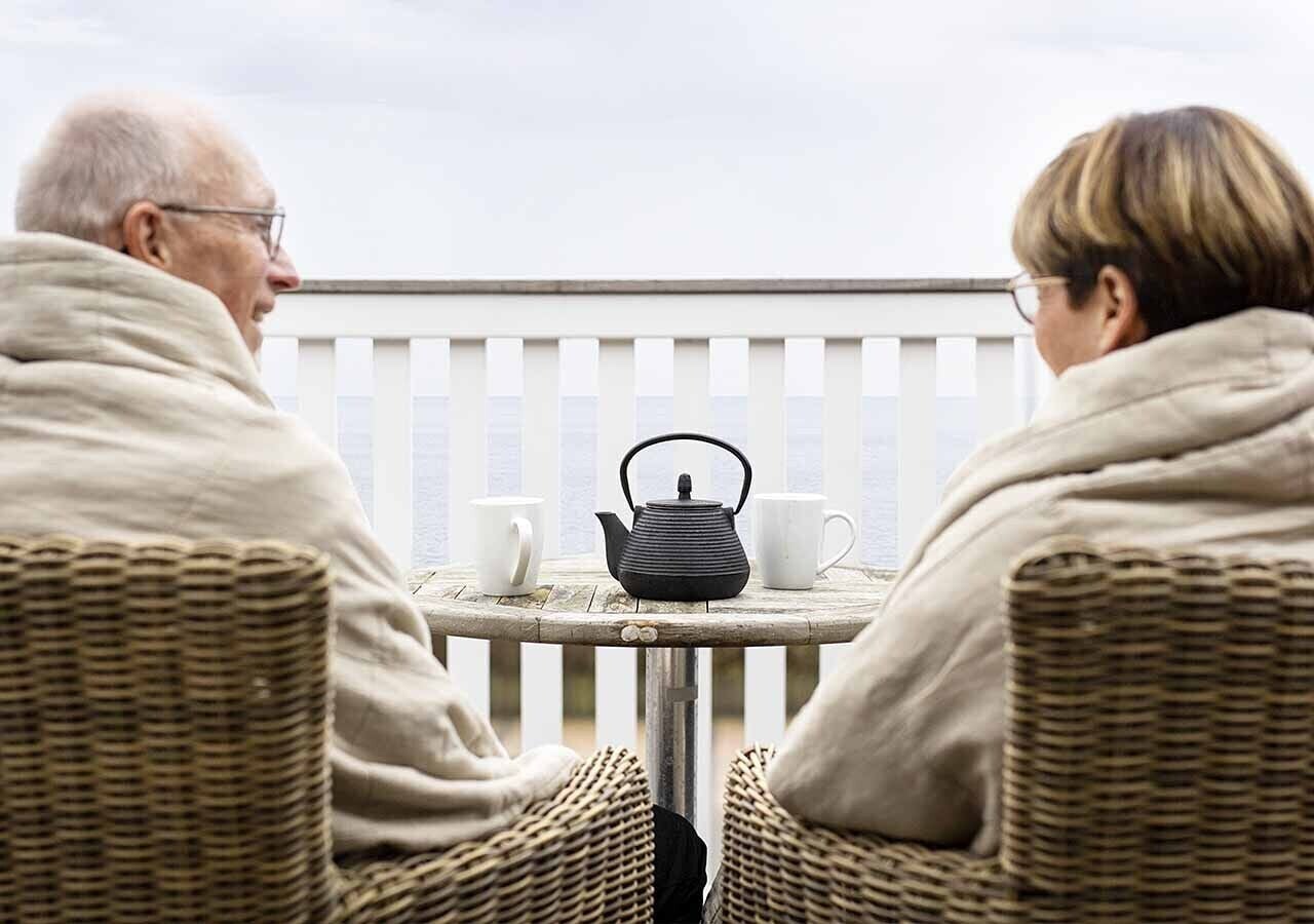 Two people seated at a round table with a kettle and two cups, on a balcony with a railing.