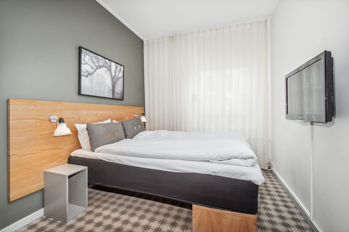 A modern hotel room with a bed, a bedside table, a wall-mounted TV, and a window with curtains.