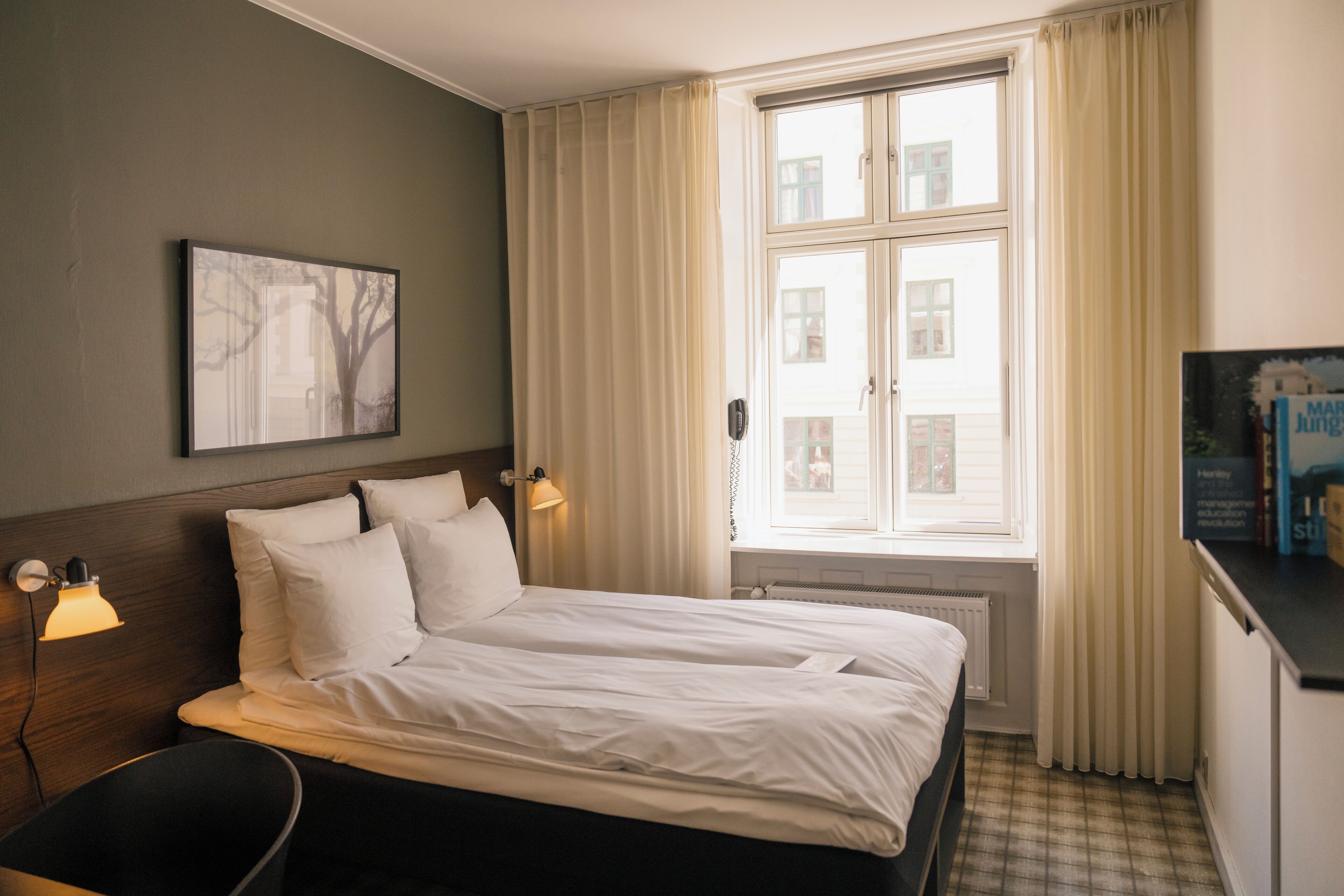 A hotel room with a bed, bedside lamps, a window with curtains, a radiator, and a wall-mounted picture.