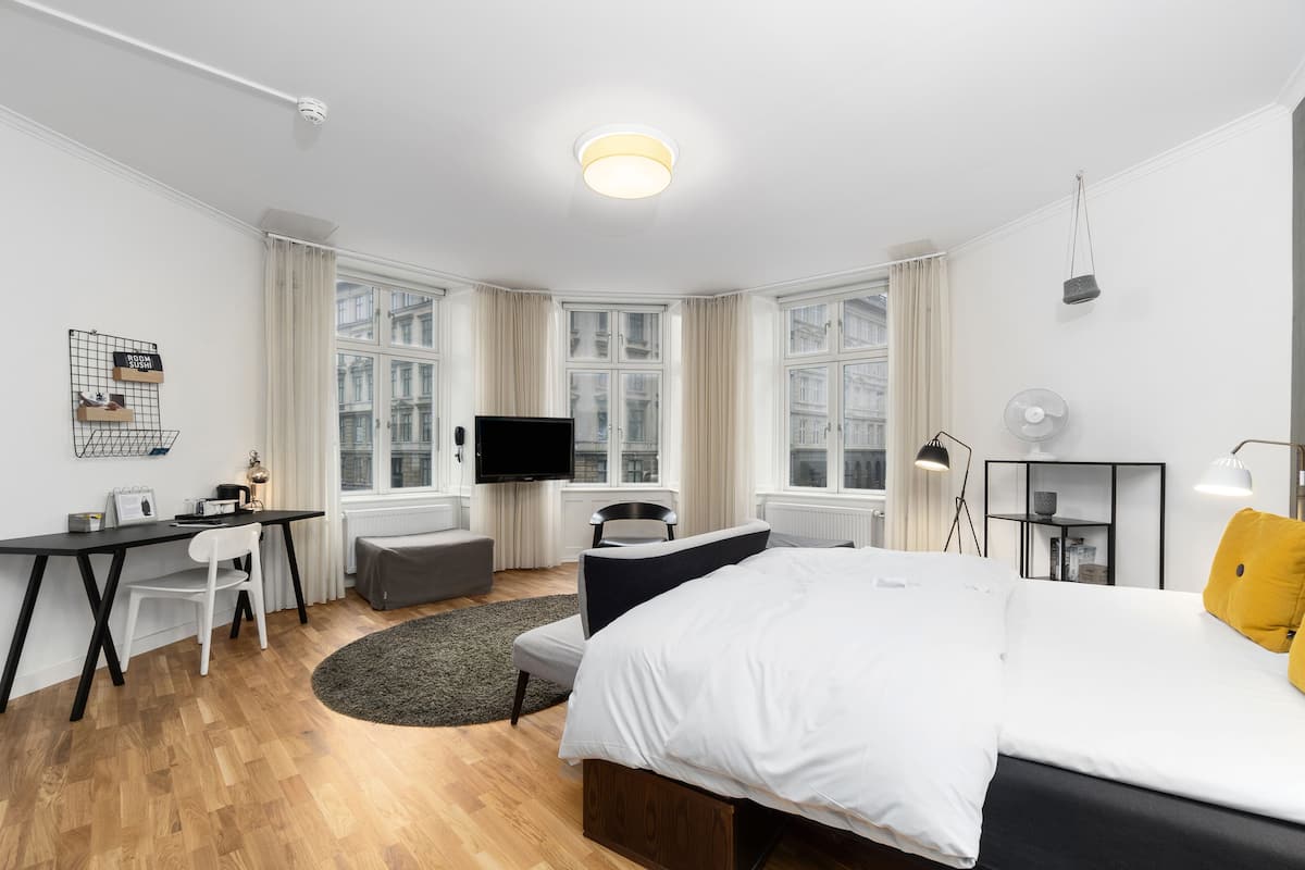 A modern hotel room with a large bed, a desk with a chair, a television, and a view of the city through large windows.