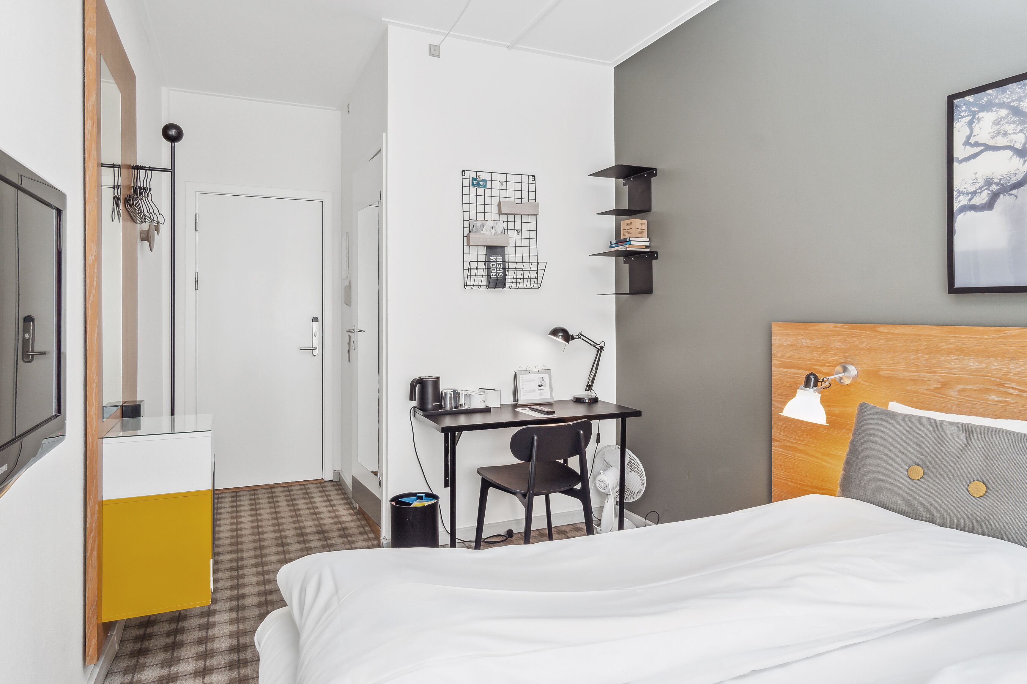 A hotel room with a bed, a desk, a chair, a desk lamp, a wall-mounted shelf, and a trash bin.