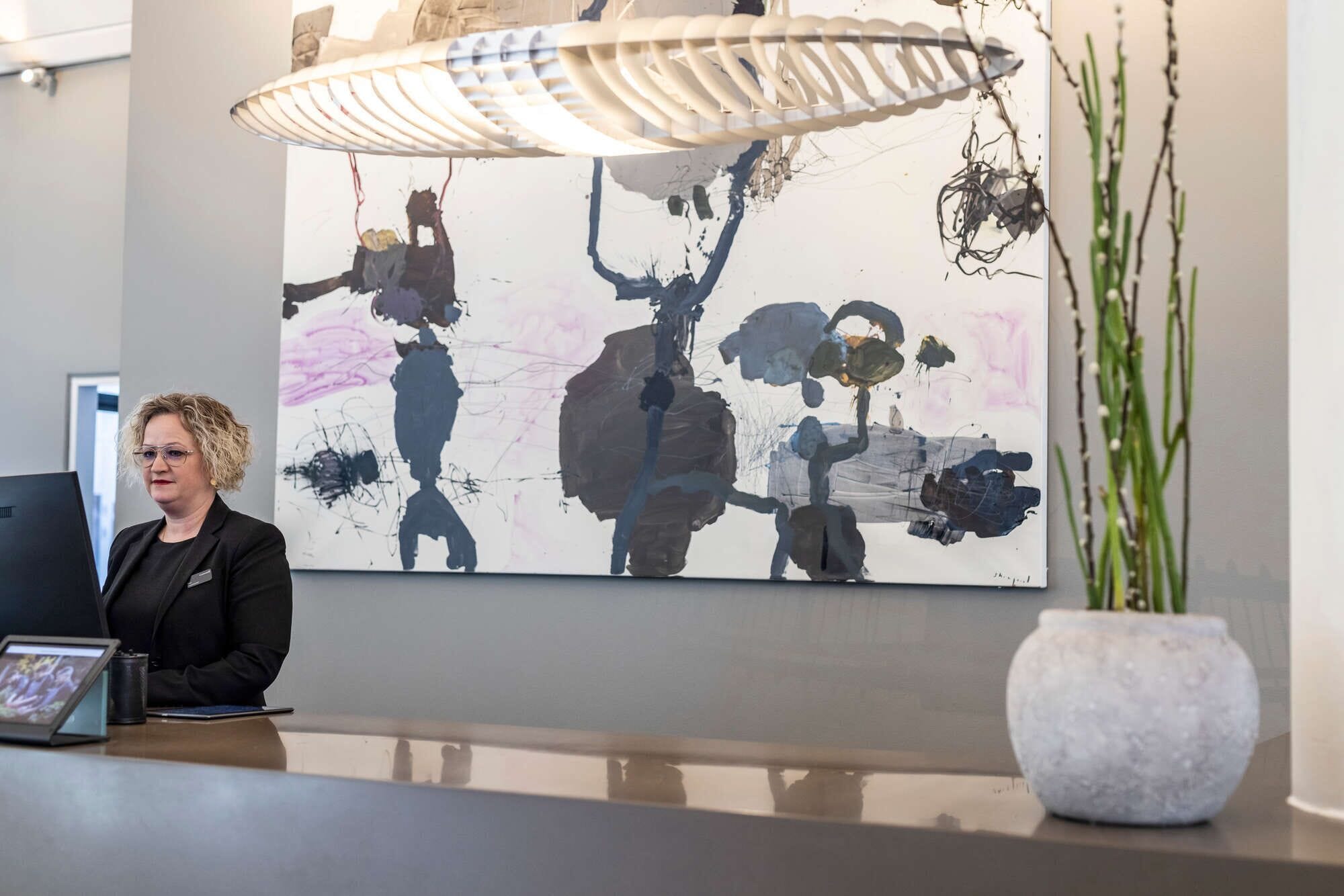 A reception desk with a large abstract painting, a potted plant, and a modern pendant light.