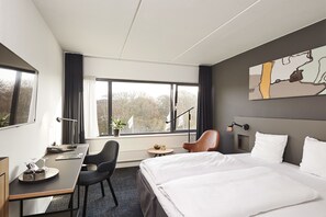 A hotel room with a bed, desk, chair, and a large window offering a view of trees. - Comwell Kolding (Kolding)