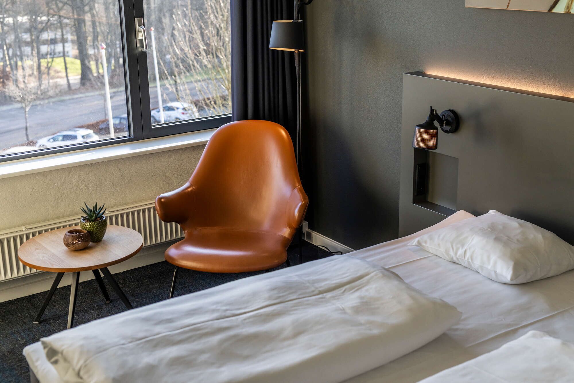 Habitación de hotel con cama, una silla, una mesita con una planta y vista a la calle por la ventana.