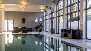 Une piscine intérieure dotée d’un sol carrelé, entourée d’espaces de détente avec des coussins et des plantes, de grandes fenêtres et d’un design moderne au niveau du plafond.