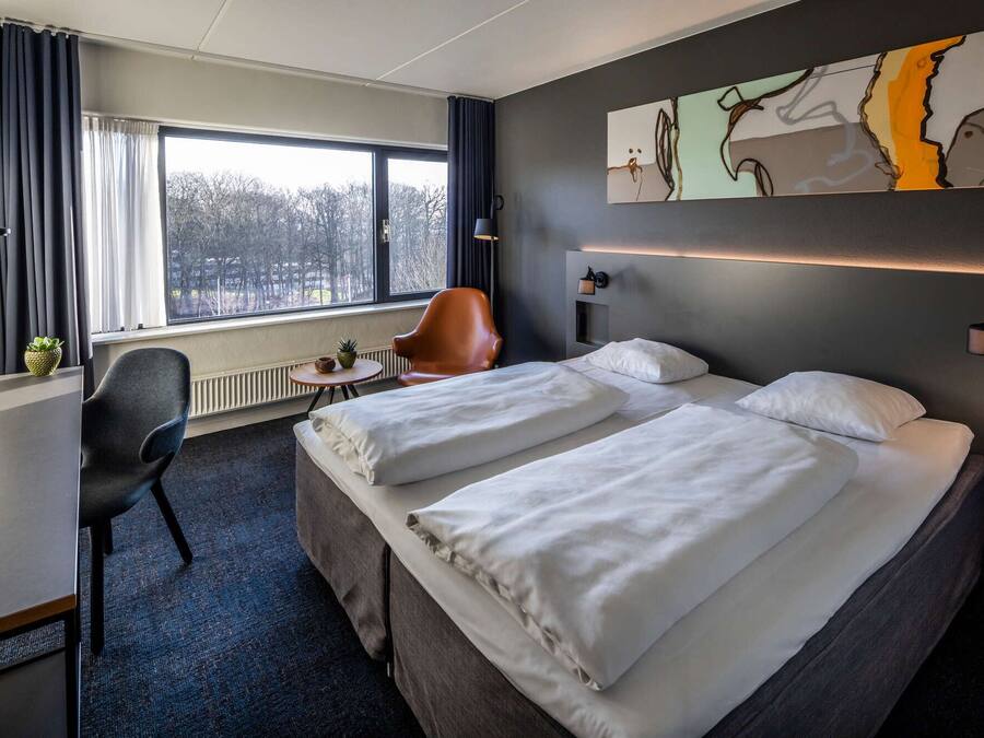 Habitación de hotel con dos camas, un escritorio, una silla, un ventanal con cortinas, una obra de arte colgada en la pared y una lámpara de noche.