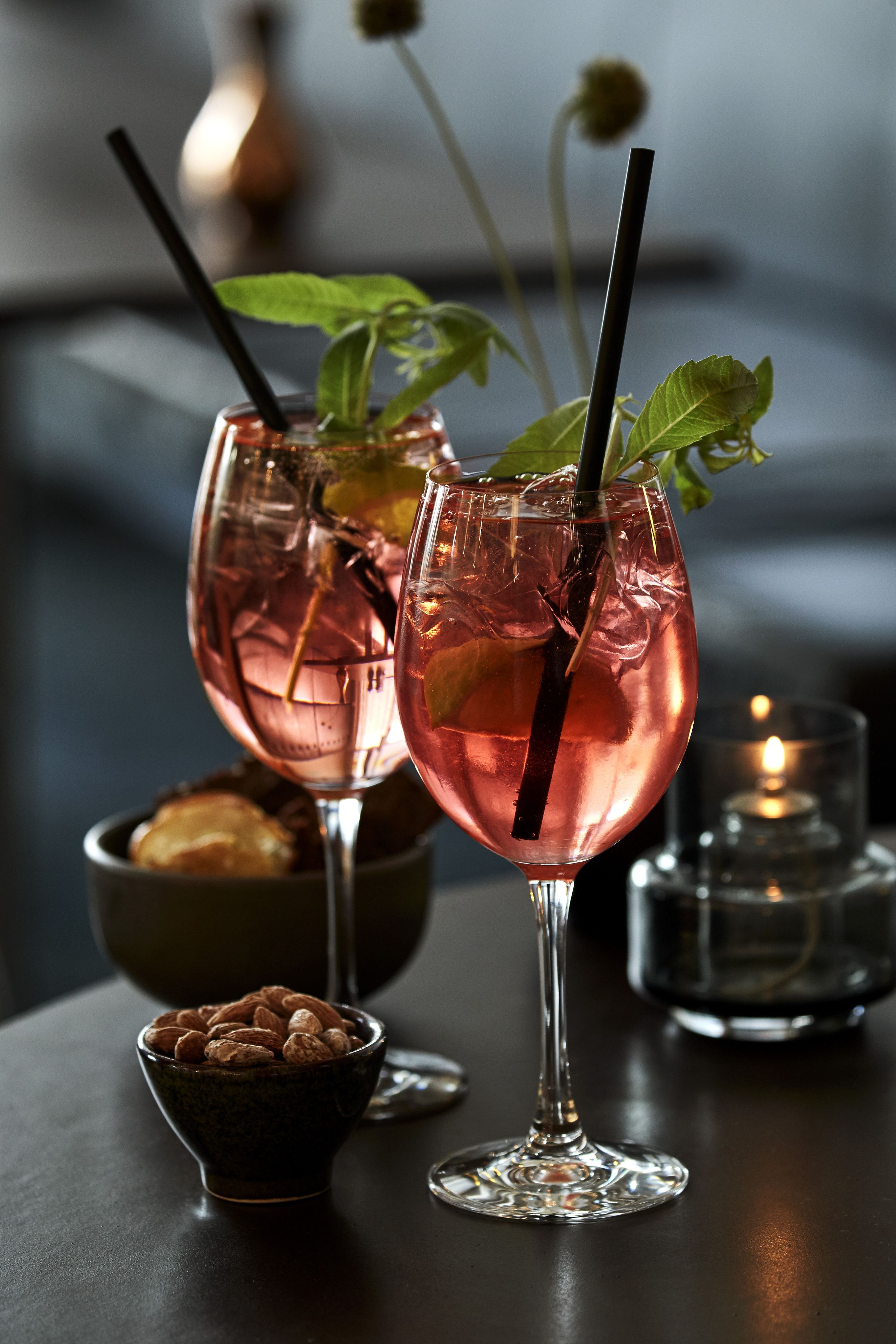 Two glasses of pink beverage with straws and mint leaves, a bowl of almonds, and a candle on a table.