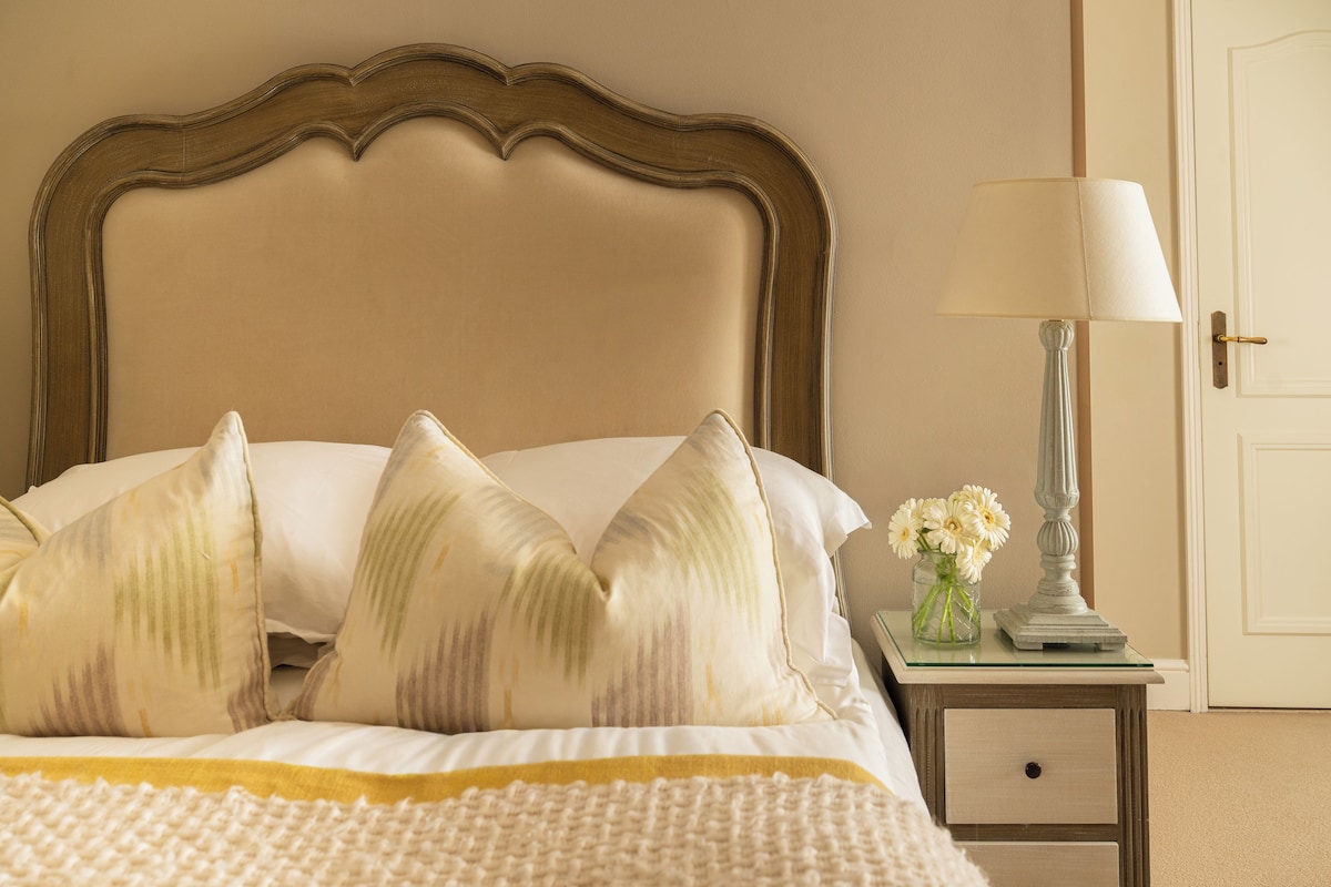 Una cama bien hecha con cabecera, una mesita de noche con lámpara y un jarrón con flores.