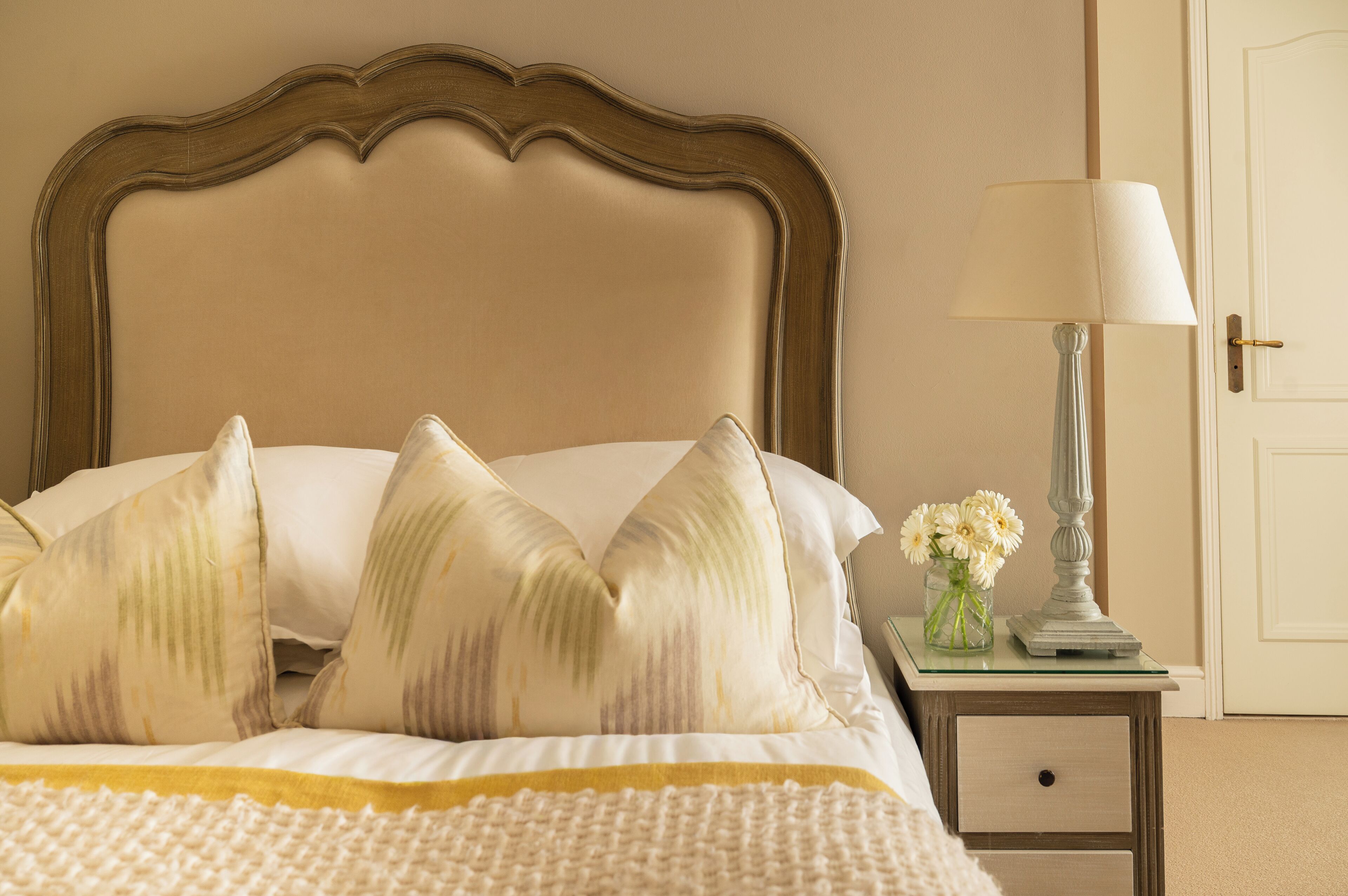 Een net opgemaakt bed met een hoofdbord, een nachtkastje met een lamp en een vaas met bloemen.