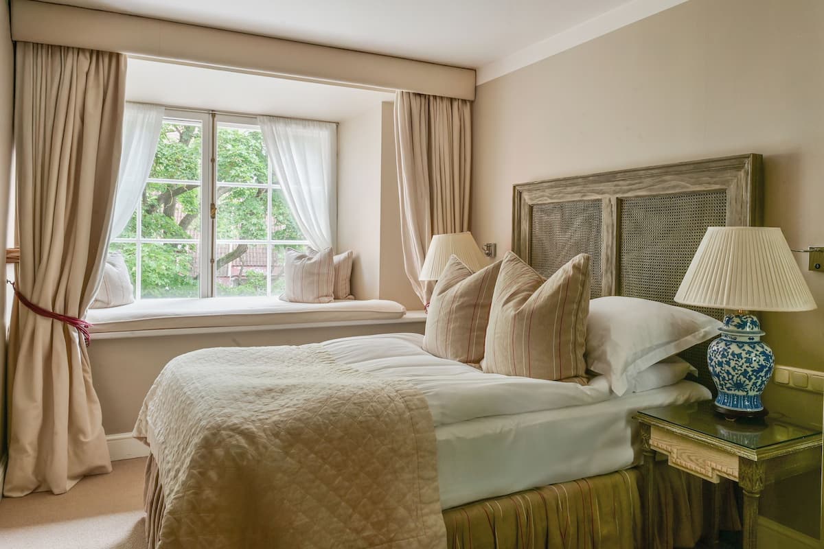 A neatly made bed with a quilted cover, a bedside lamp, and a window with curtains.