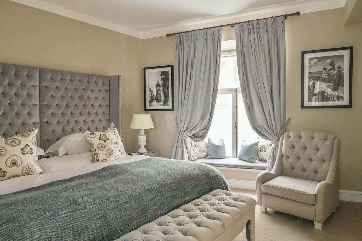 A bedroom with a tufted headboard, a large bed with patterned pillows, a grey throw blanket, a bedside lamp, a window seat with curtains, and a chair.