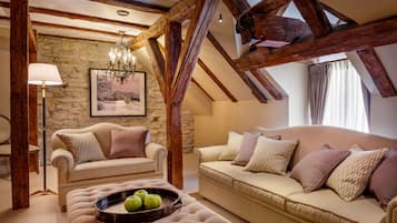 A cozy living room with a stone wall, wooden beams, a chandelier, a sofa, a chair, and a table with apples.