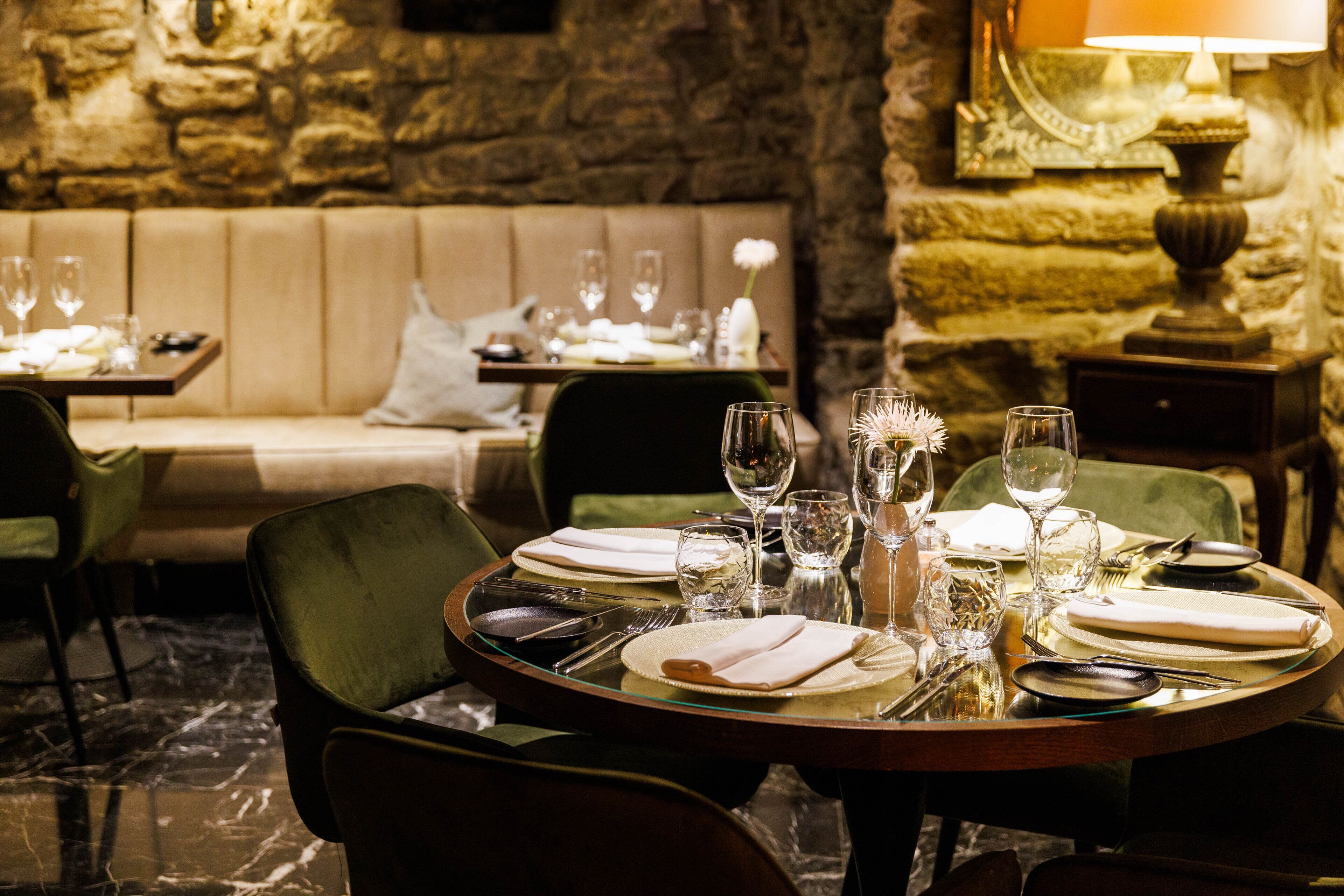 Un comedor con una mesa redonda dispuesta para dos personas, con platos, cubiertos y copas de vino. Un sofá y sillones se encuentran en el fondo, y una pared de piedra complementa el ambiente.