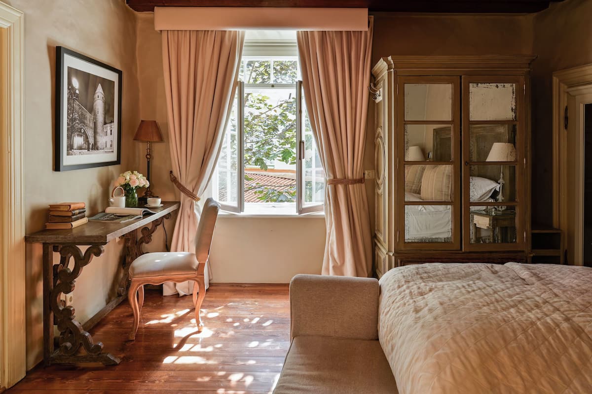 A bedroom with a bed, a desk with books and a lamp, a chair, and a window with curtains.